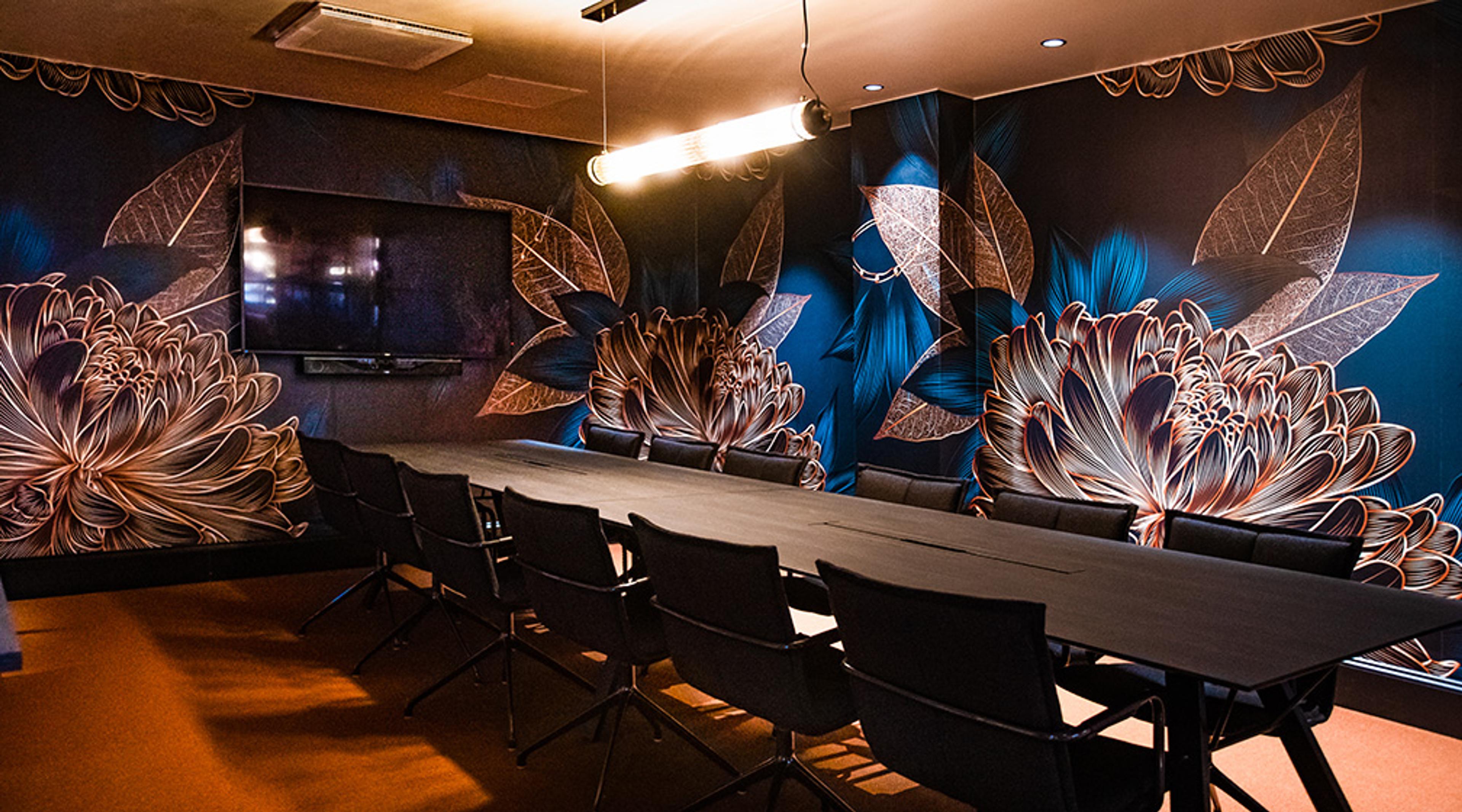 Conference room with long table, black chairs and decorative floral and leaf patterned wall. Flatscreen television mounted on the wall.