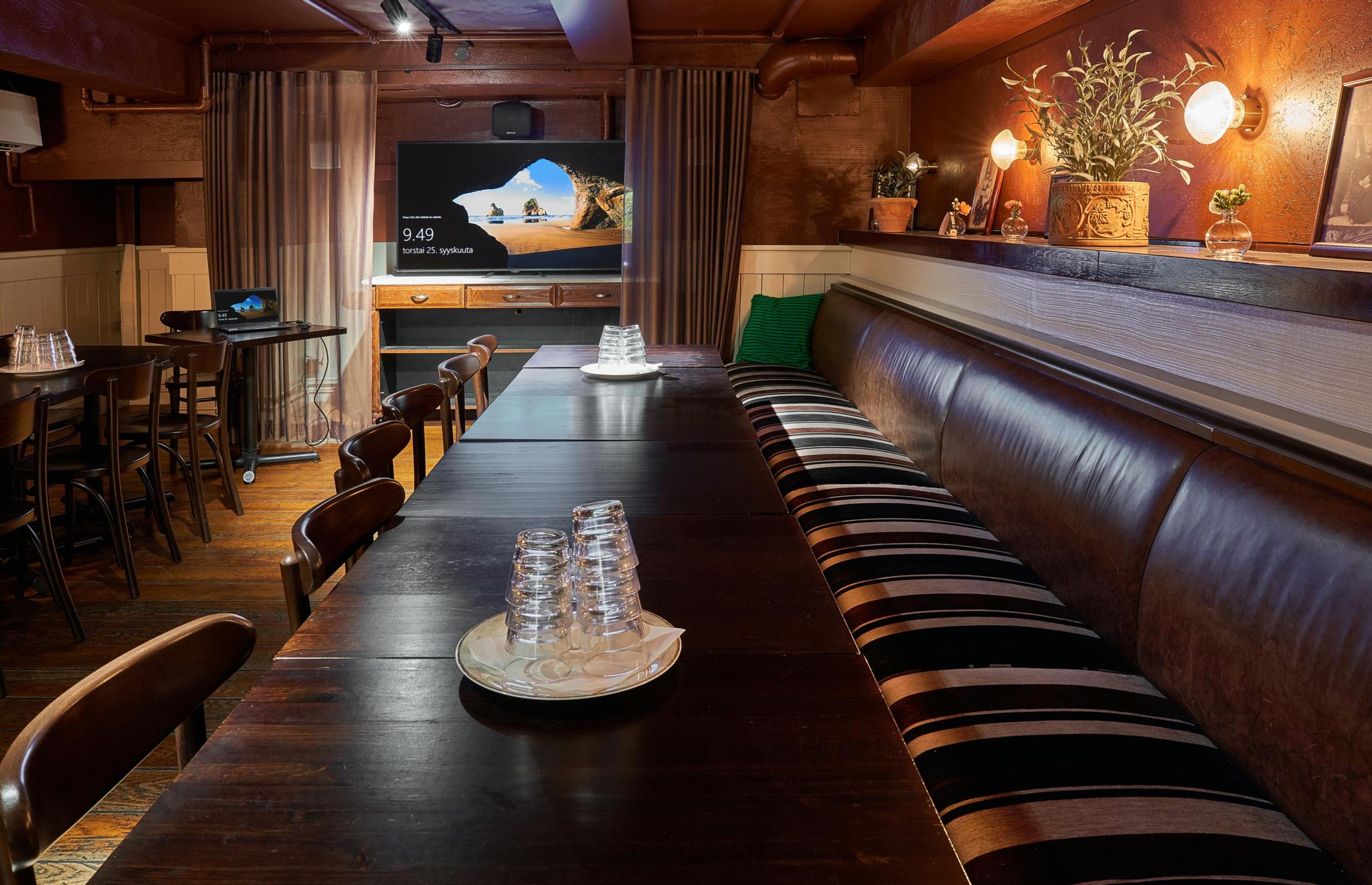 Pieni kokoustila, jossa pitkä pöytä, raidallinen sohva, puiset tuolit ja näyttö. Osittain paneloitu tila lämpimällä valaistuksella ja puulattialla.