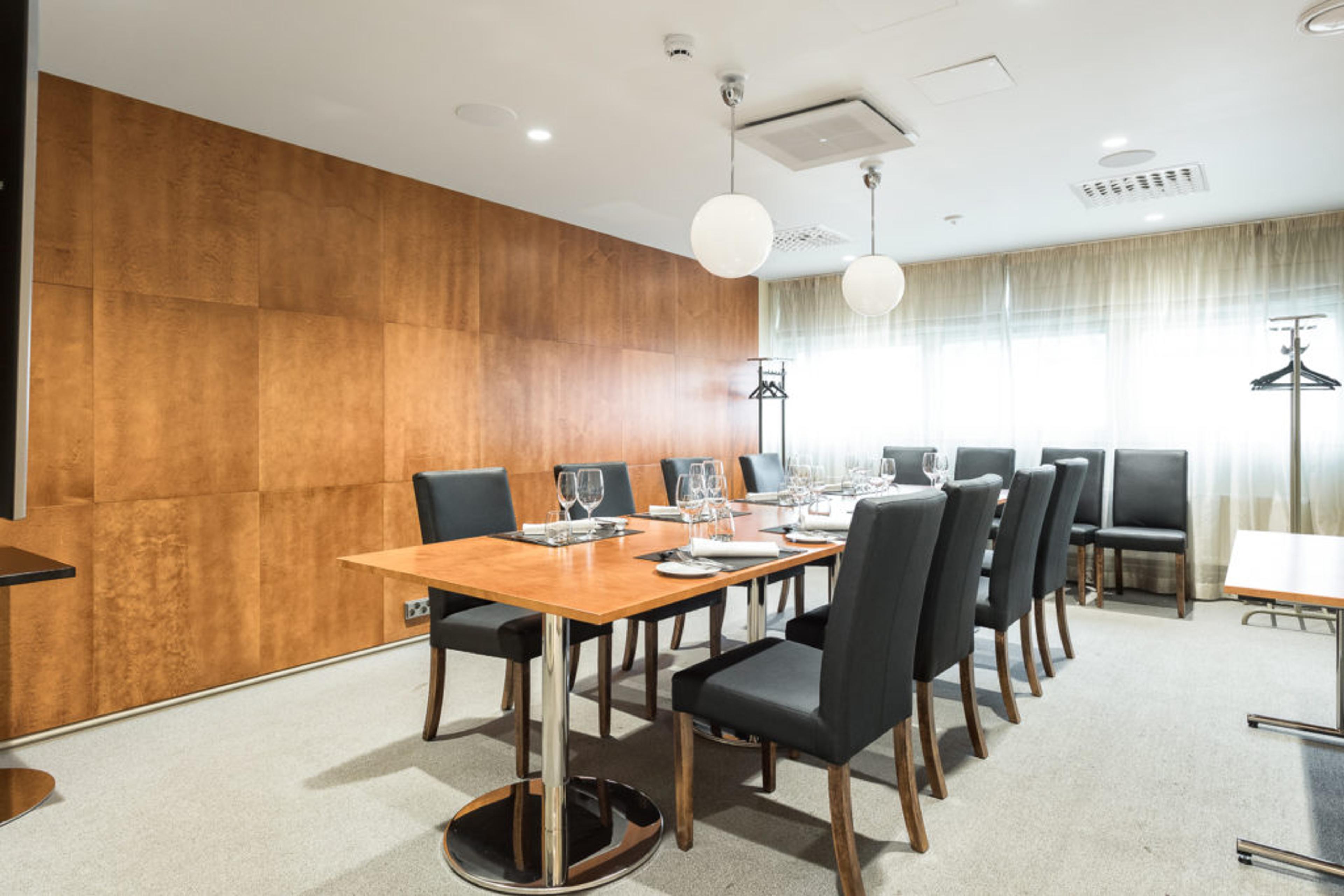 Moderni kokoushuone puupaneeliseinällä, pitkä pöytä mustilla tuoleilla, kuvassa lasit ja astiat valmiina. Vaalea lattia, pyöreät valaisimet katossa.