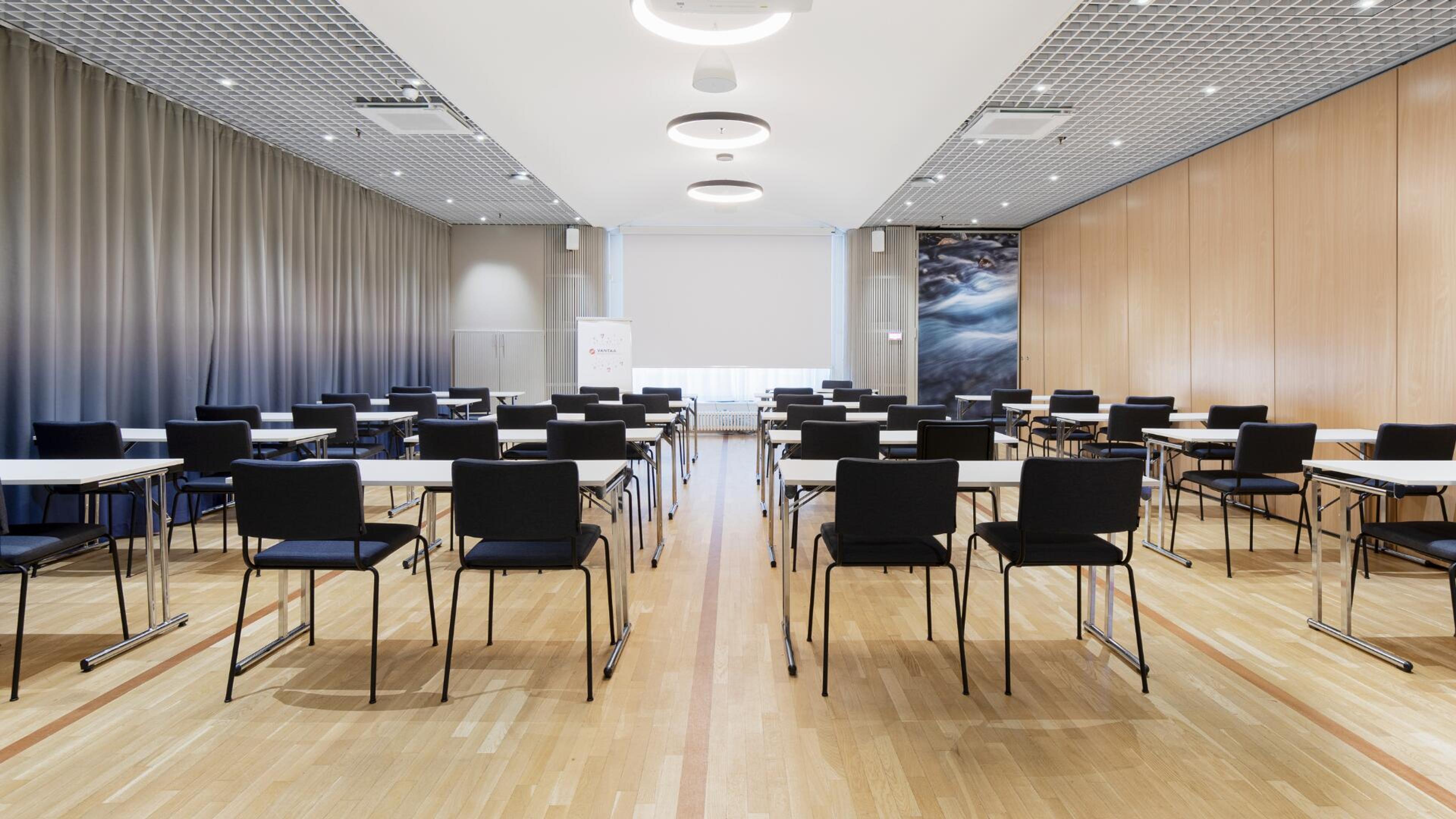 Moderni kokoushuone vaalean puulattialla, mustilla tuoleilla ja valkoisilla pöydillä. Tilassa on harmaat verhot ja puupaneeliseinä.