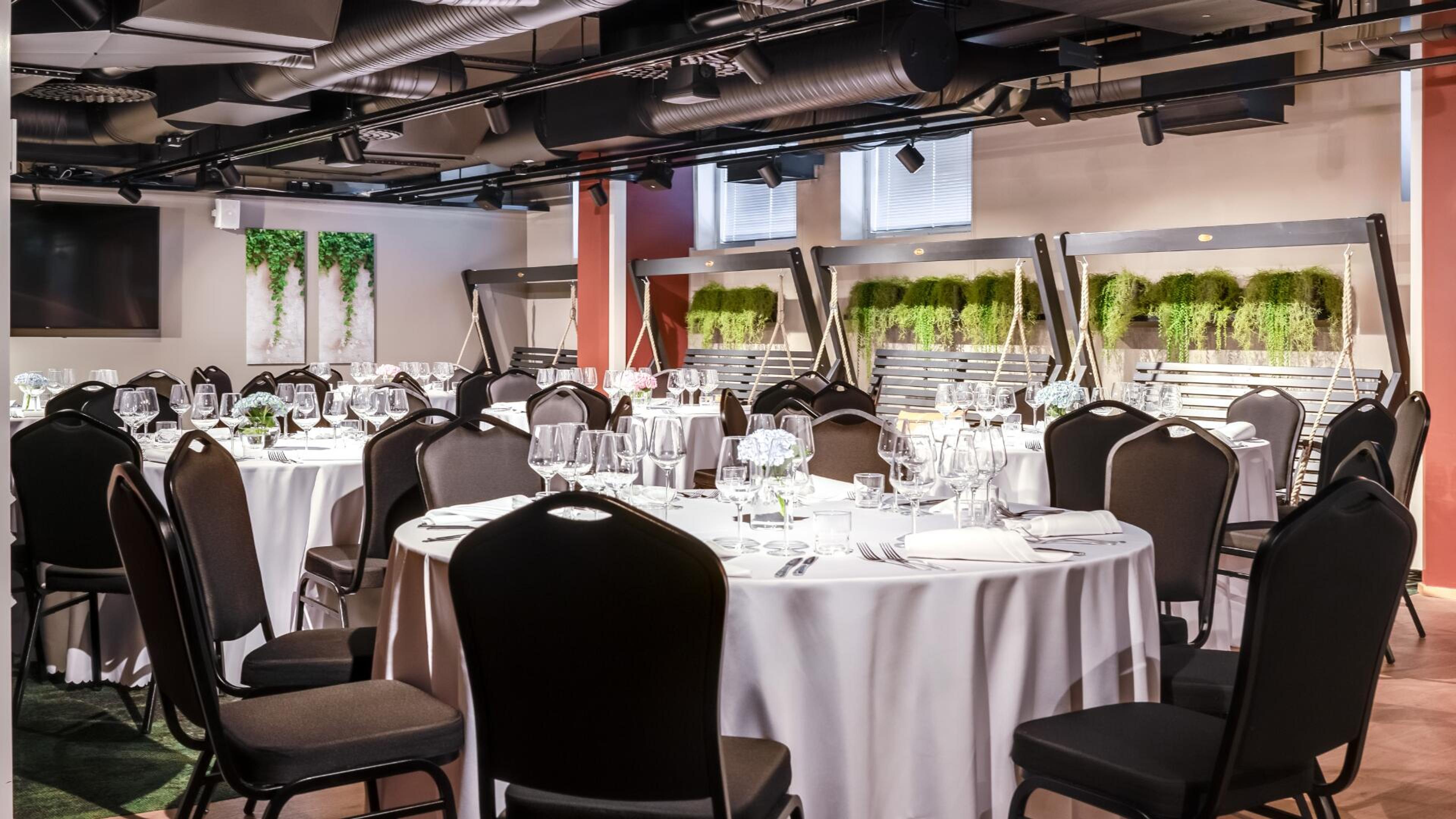 Juhlatila, jossa valkoliinaiset pyöreät pöydät ja tummat tuolit. Seinällä viherkasveja ja riippuvia istutuksia.