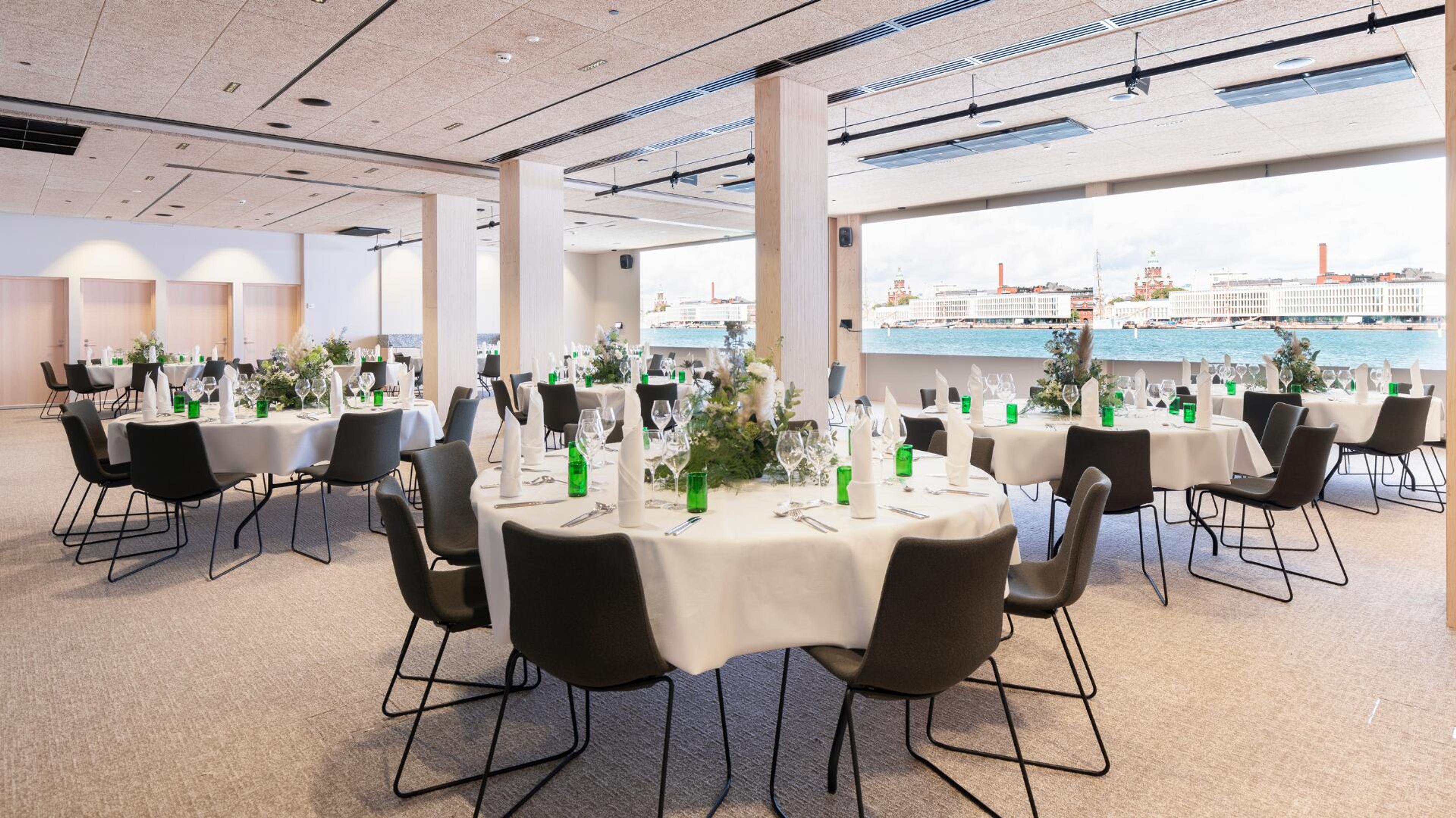 Valoisa ravintolasali, jossa pyöreitä pöytiä valkoisin liinoin ja mustia tuoleja. Suurista ikkunoista avautuu merinäkymä kaupungin siluetin edustalla.