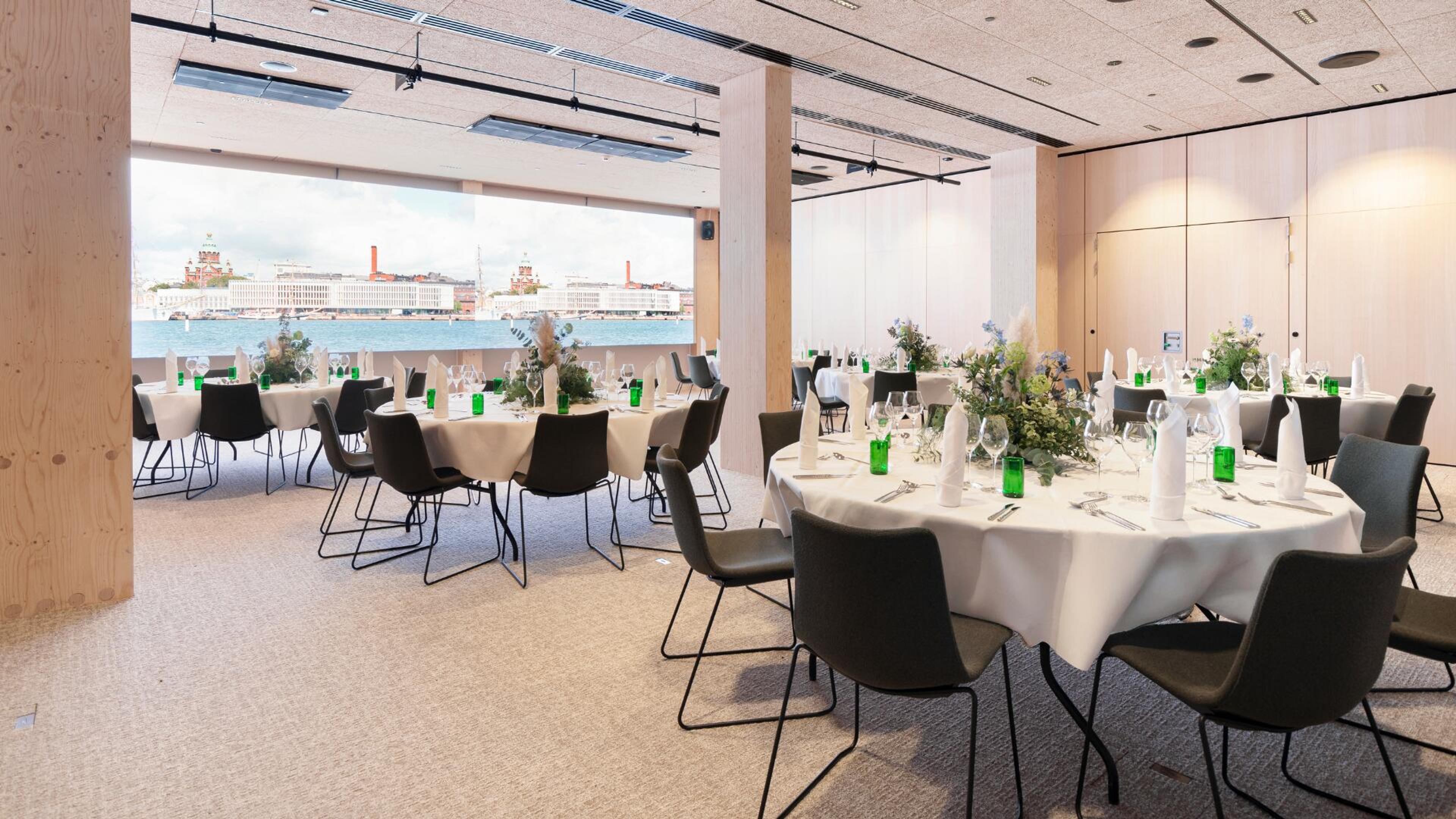 Ravintolasali pyöreillä pöydillä, koristeellisin kukka-asetelmin ja vihreillä pulloilla. Suurista ikkunoista näkyy kaupungin siluetti veden takana.