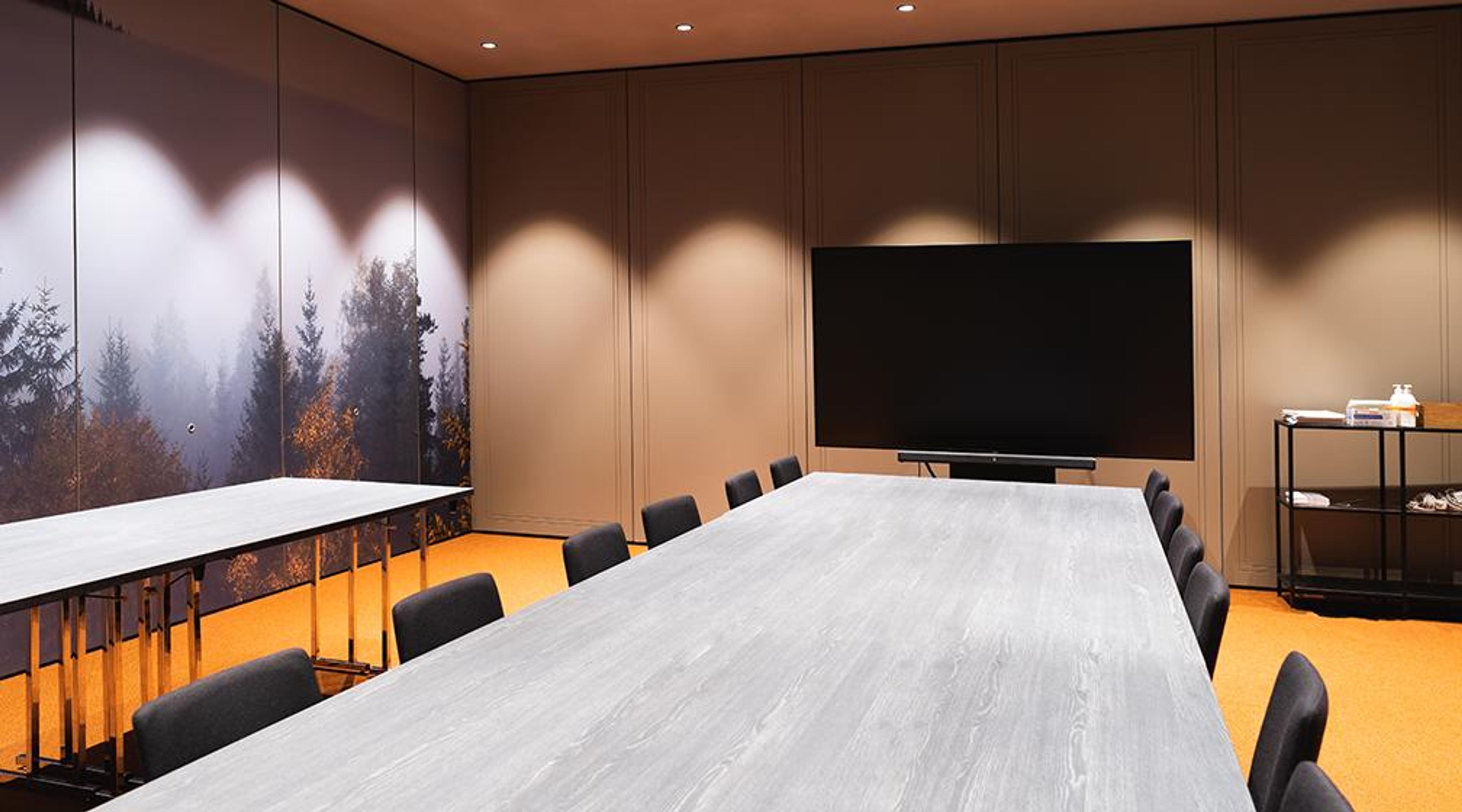 Modern conference room with long grey table and black chairs, orange floor, forest landscape wall decor, and a TV screen mounted on the wall.