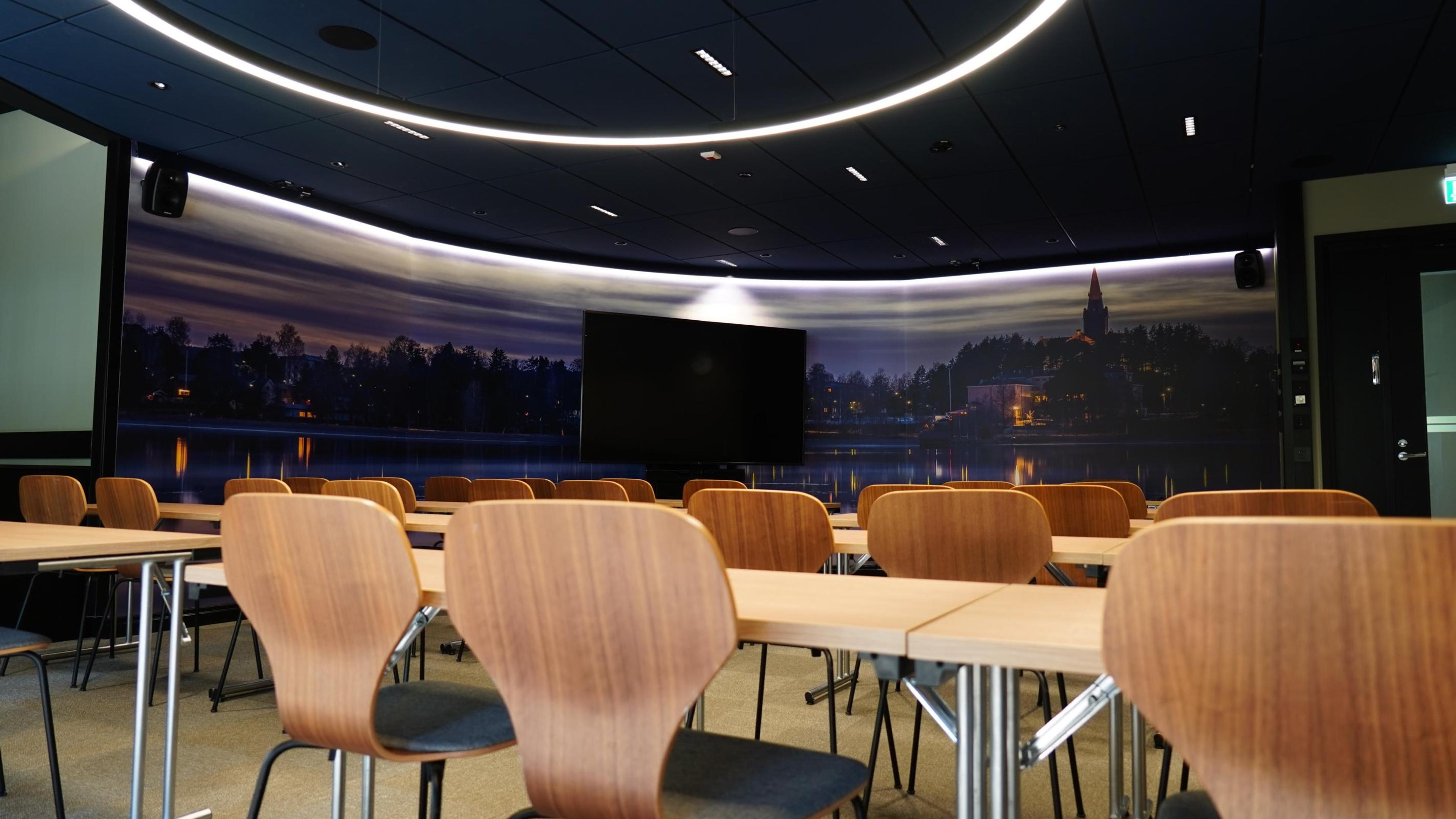 Mötesrum med trästolar och bord. Panoramaskärm på väggen visar kvällsvy med sjö och kyrka. Rummet har plant golv och kurvade taklampor.