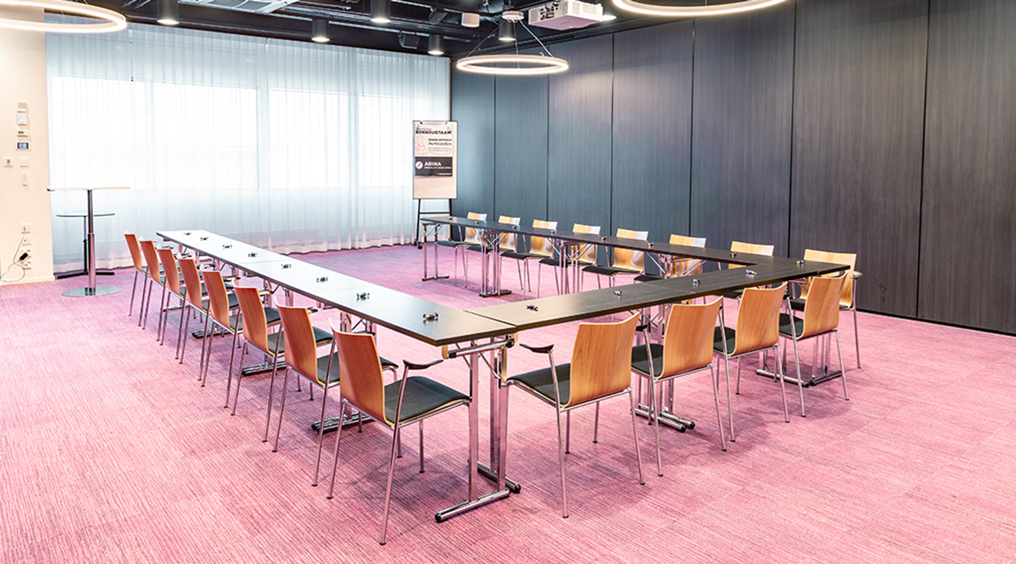 Moderni kokoustila, jossa neliön muotoinen pöytäasetelma puisilla tuoleilla. Huoneessa on vaaleanpunainen kokolattiamatto ja harmaat seinäpaneelit.