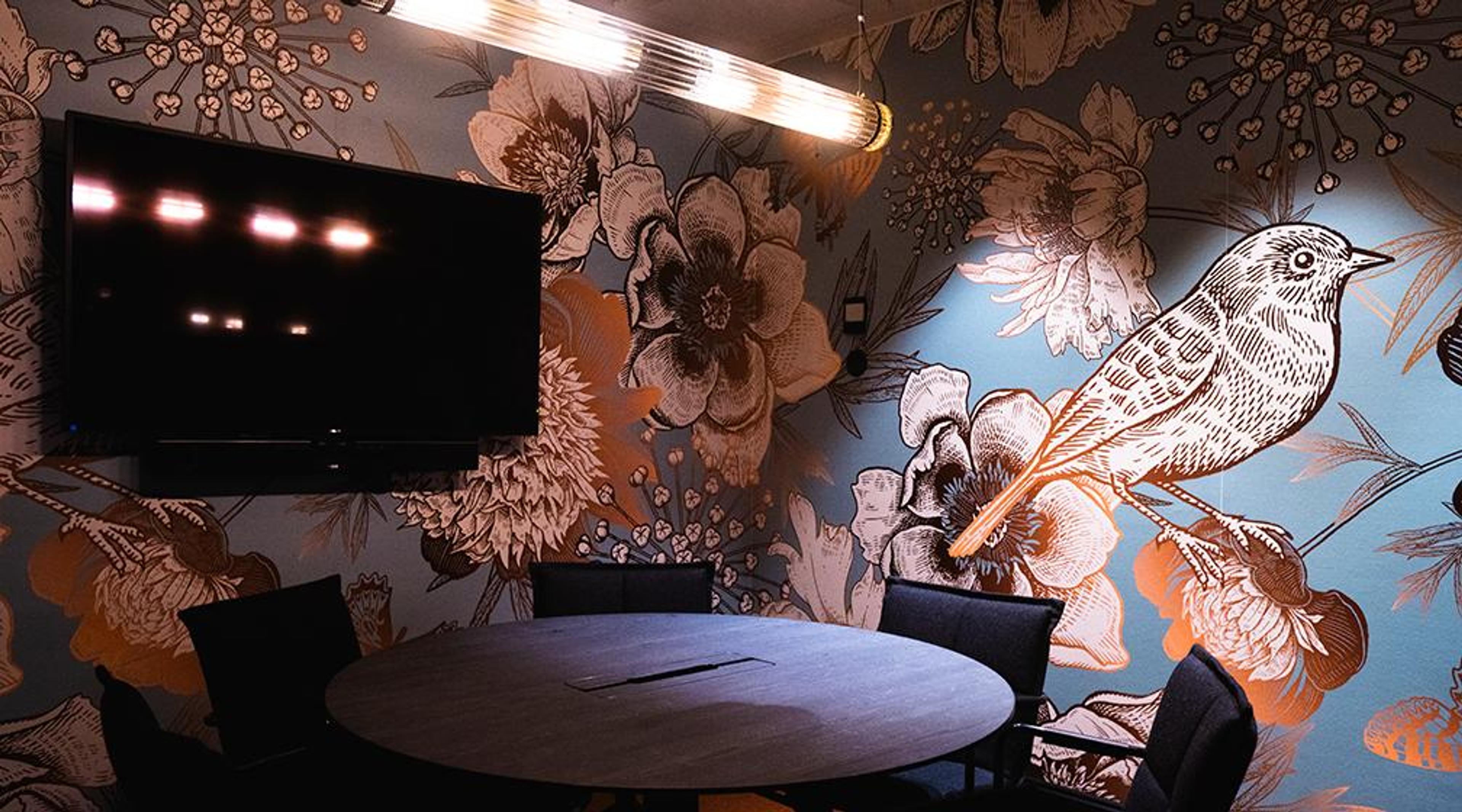 Meeting room with a round table, black chairs and a screen. Walls feature striking floral and bird patterned wallpaper.