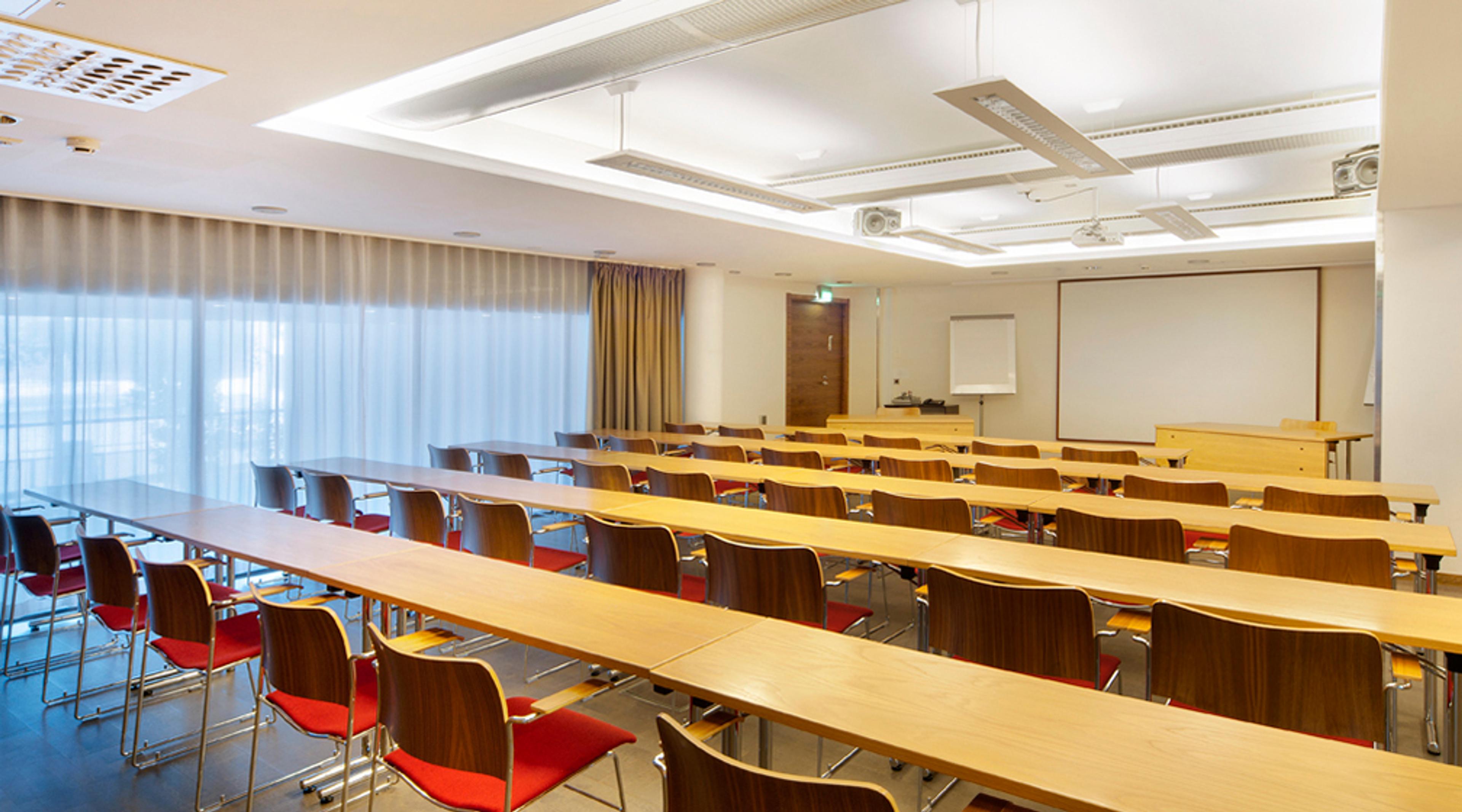 Bright meeting room with long wooden tables and red chairs arranged in rows. A white presentation board at front and large windows with curtains on one side.