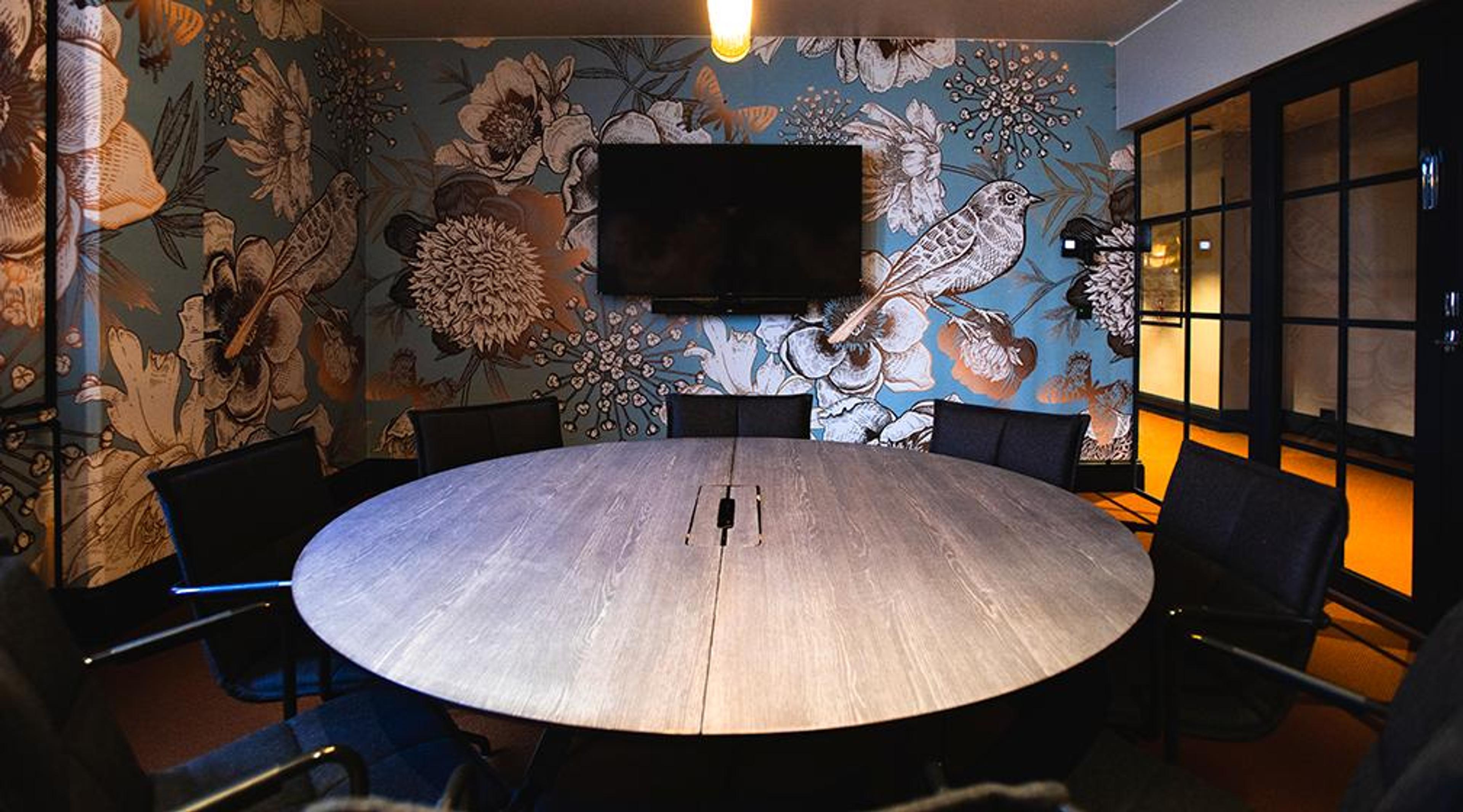 Meeting room with a round wooden table, black chairs and decorative floral and bird wallpaper. A TV screen is mounted on the wall.