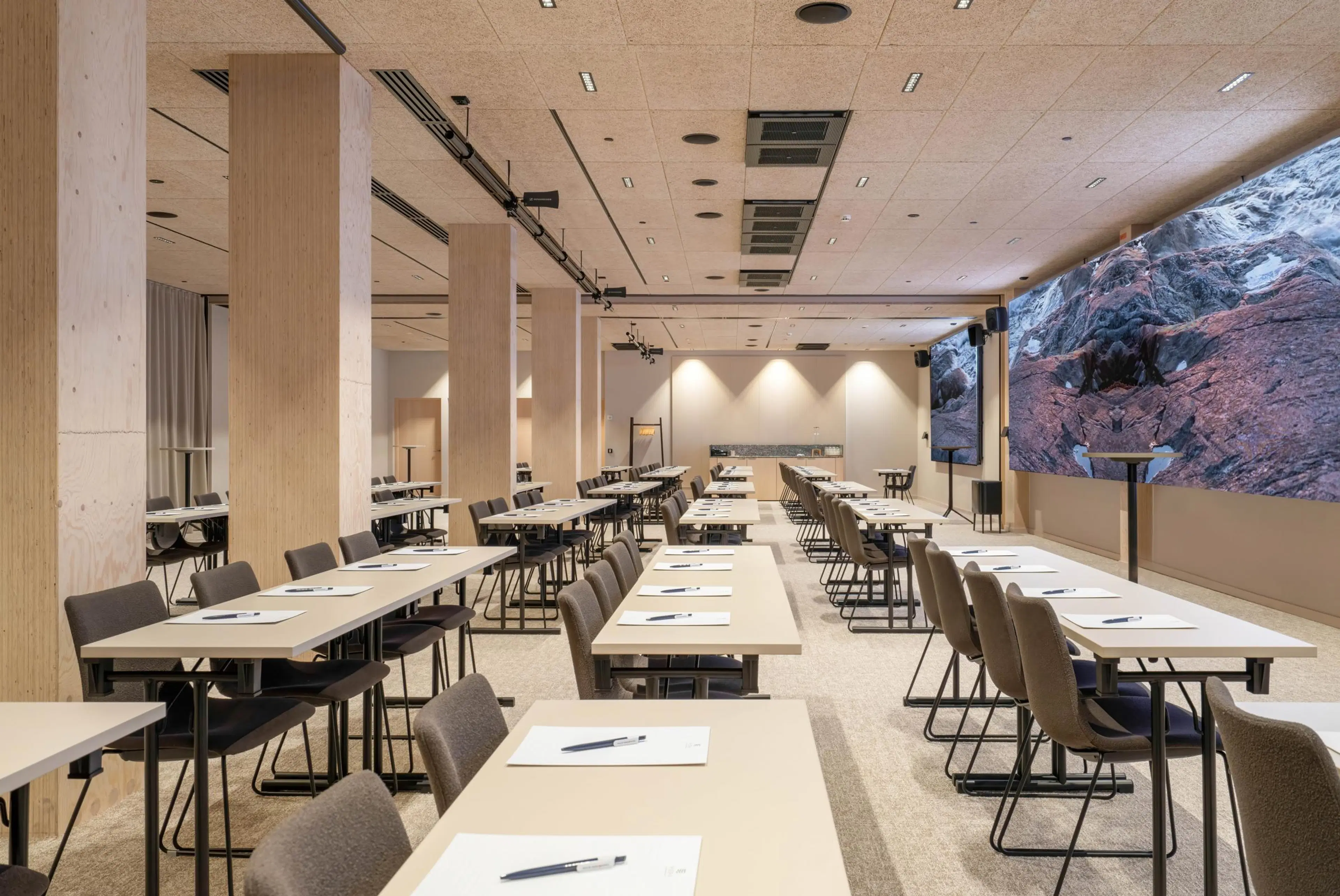Modern conference room with wooden tables arranged classroom-style, gray chairs, light-colored flooring, and a striking rock formation image on the wall. The lighting is bright and even.