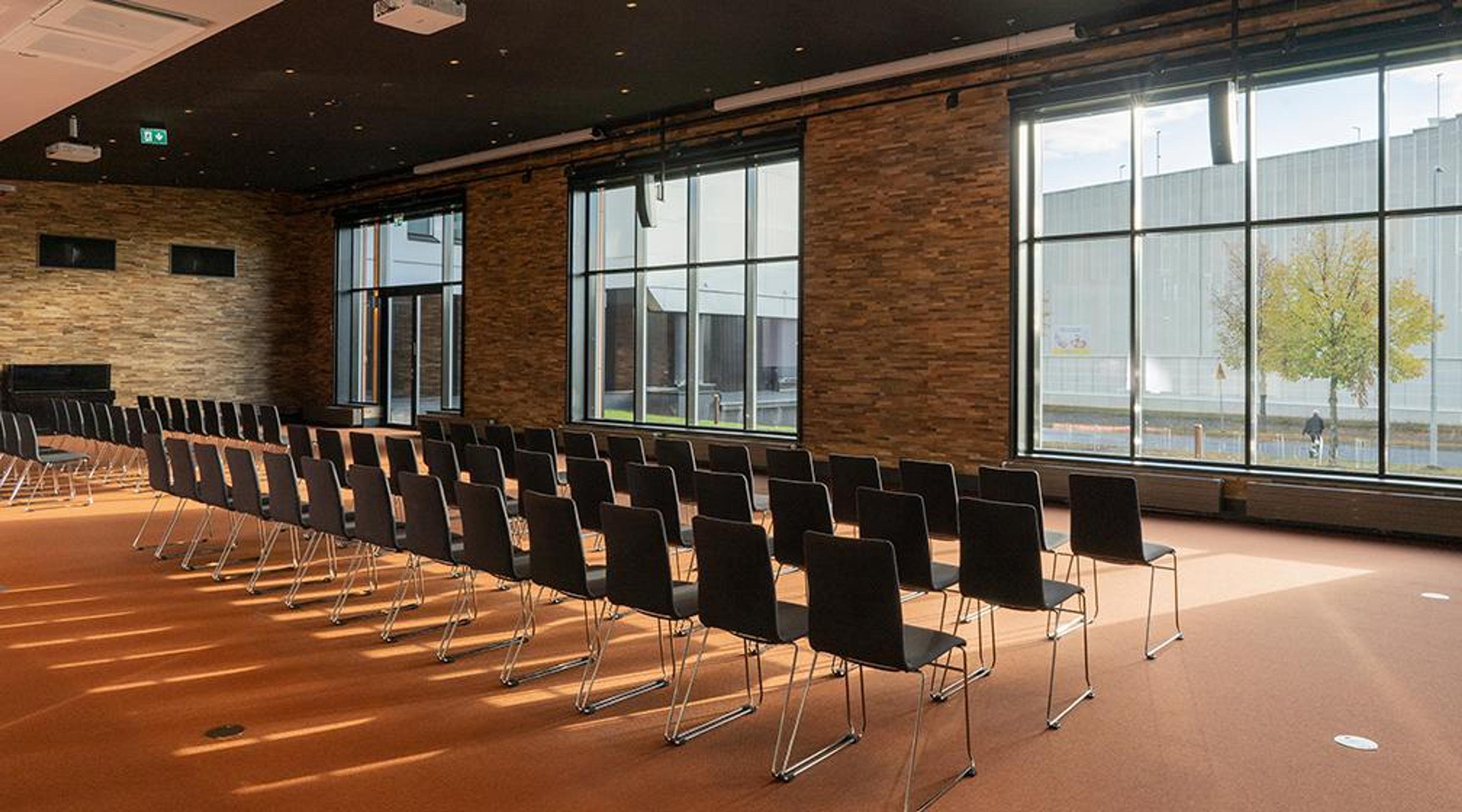 Modern auditorium with rows of black chairs on light flooring. Brick walls and large windows allowing natural light into the space.