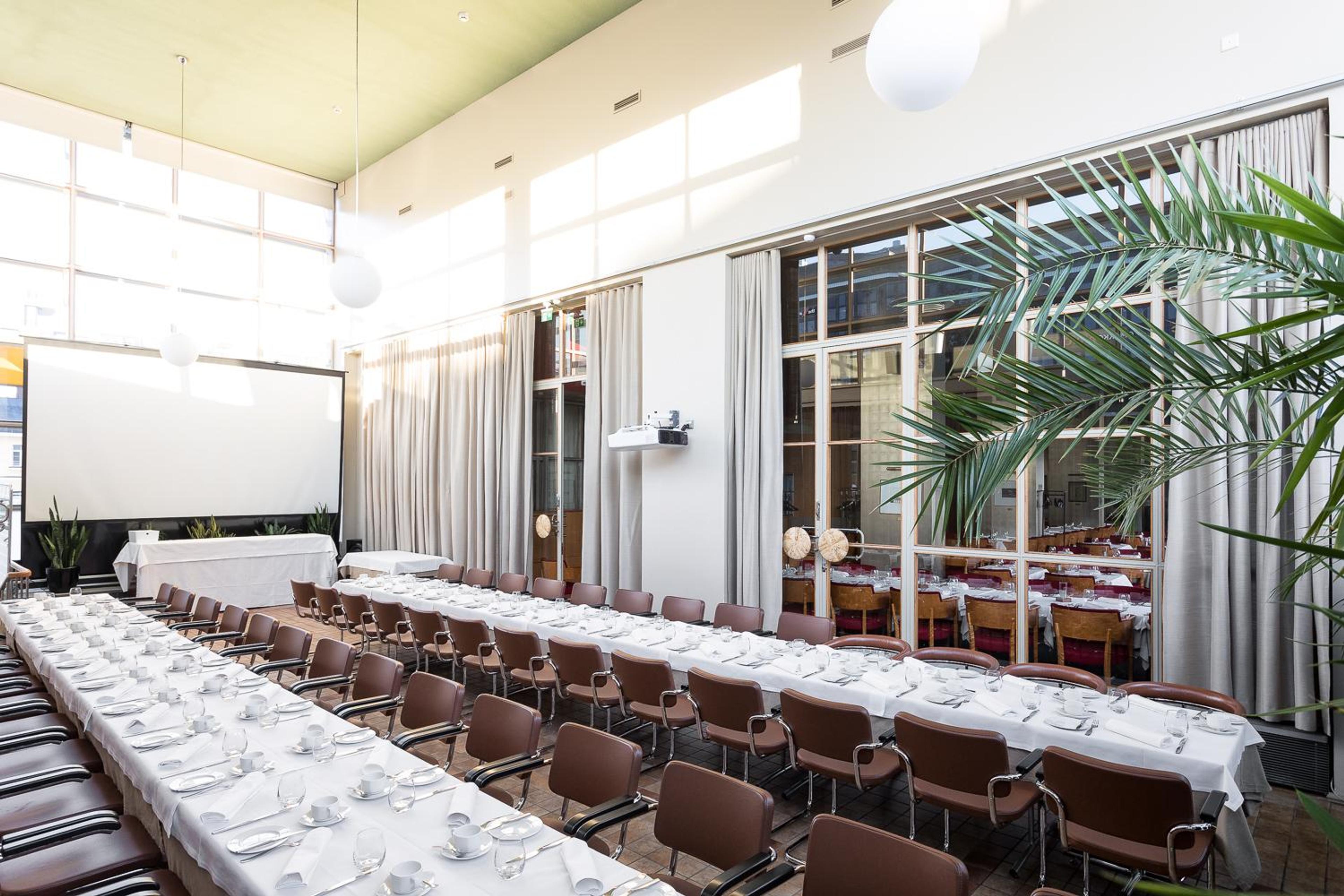 Valoisa kokoustila, jossa pitkät pöydät valkoisilla liinoilla ja kattauksilla. Ruskeat tuolit, valkokangas ja suuret ikkunat. Nurkassa vihreä palmukasvi.