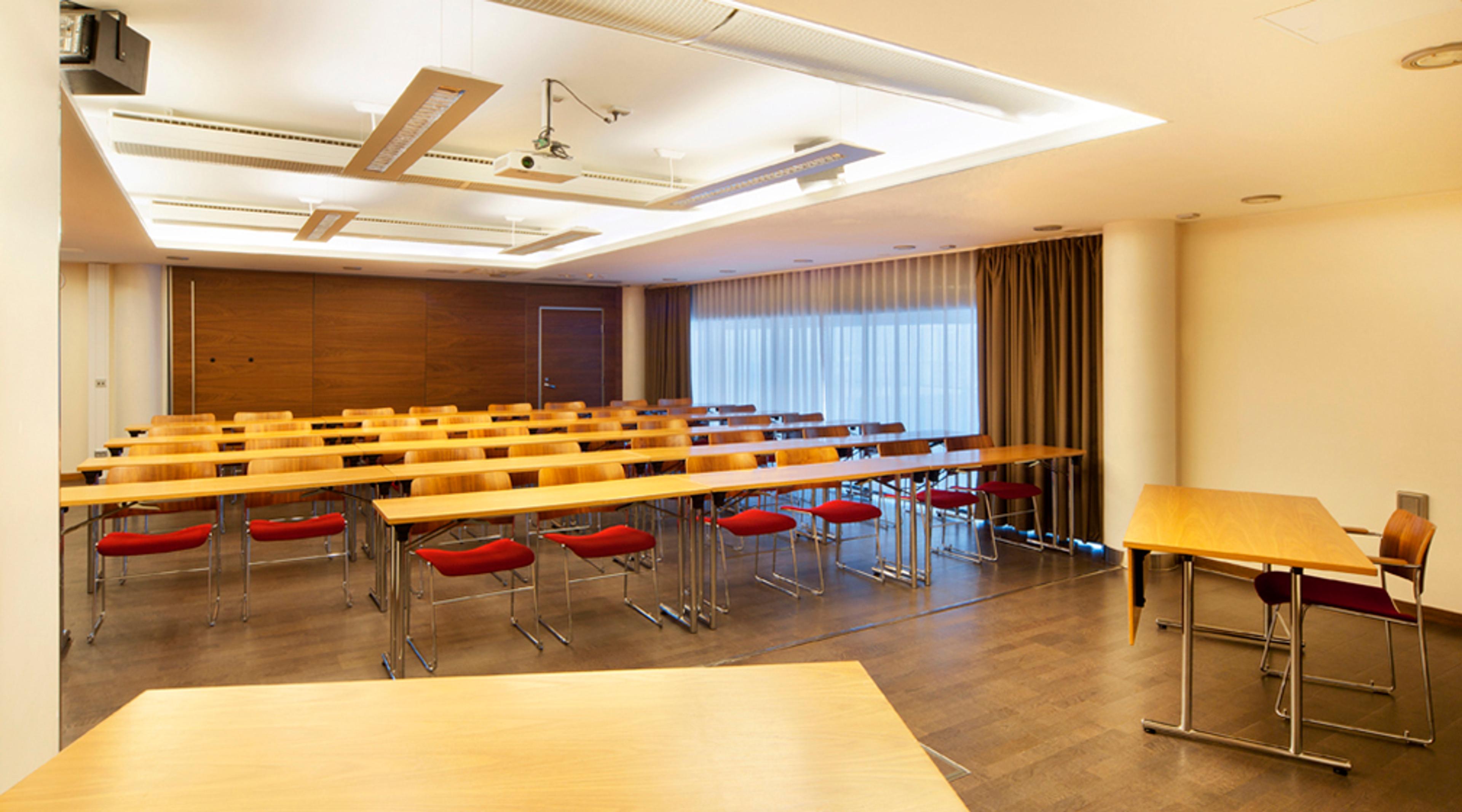 Conference room with tables arranged in rows and red chairs. Flat wooden floor, ceiling-mounted projector and large curtains on windows.