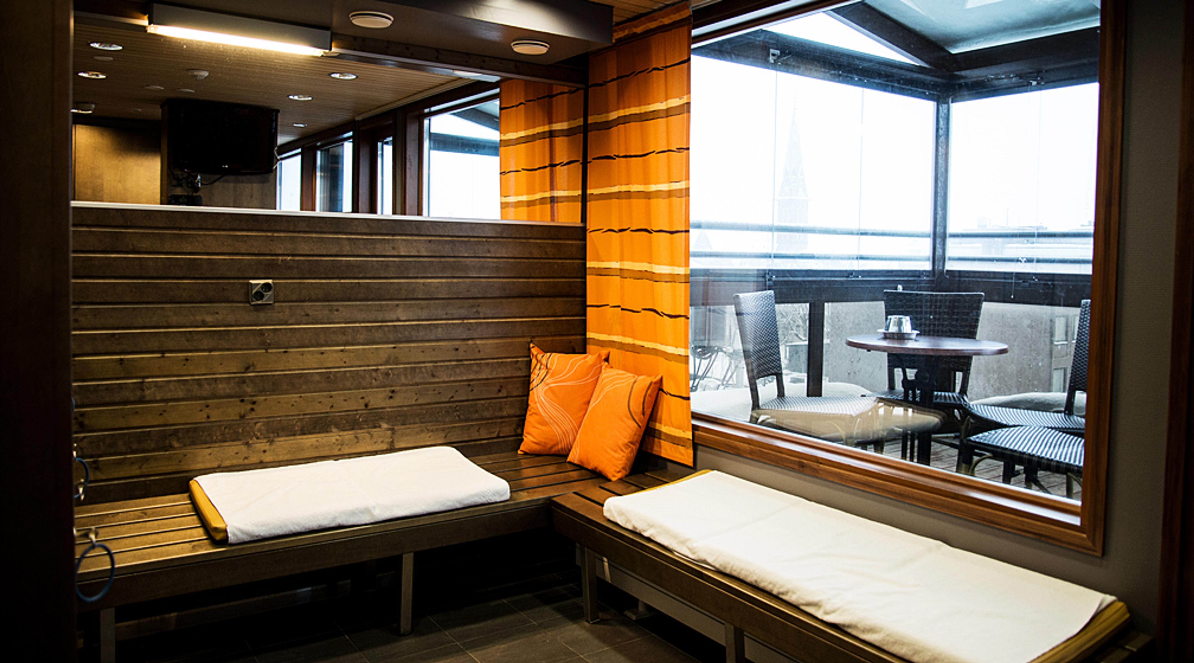Sauna, jossa puinen seinäpanelointi, oranssit tyynyt ja valkoiset pyyhkeet. Ikkunan takana näkyy ulkoterassi pöydillä.