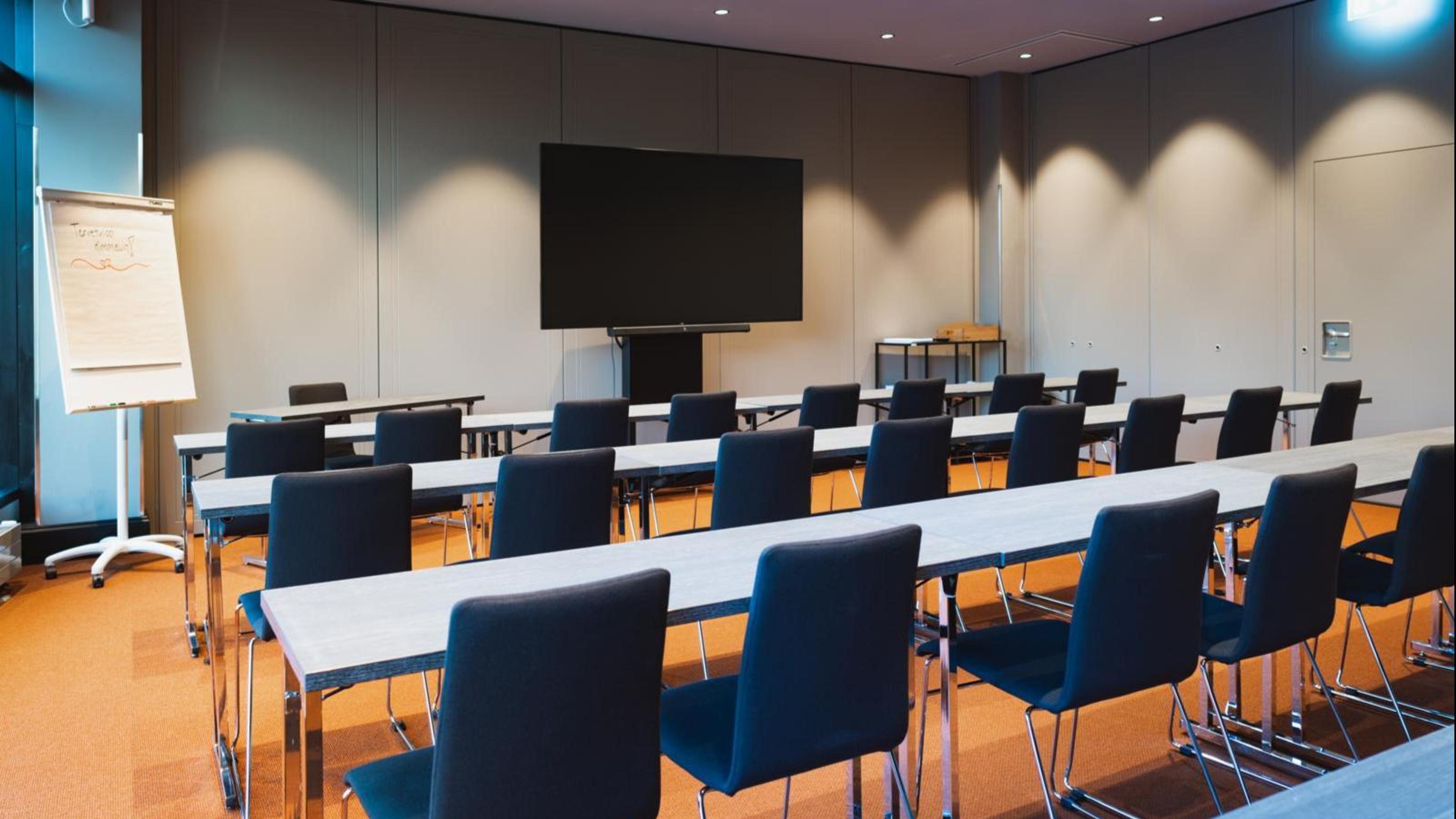 Meeting room with long tables and blue chairs arranged in rows, large display screen on wall and flipchart on left. Orange carpeted floor and even lighting.