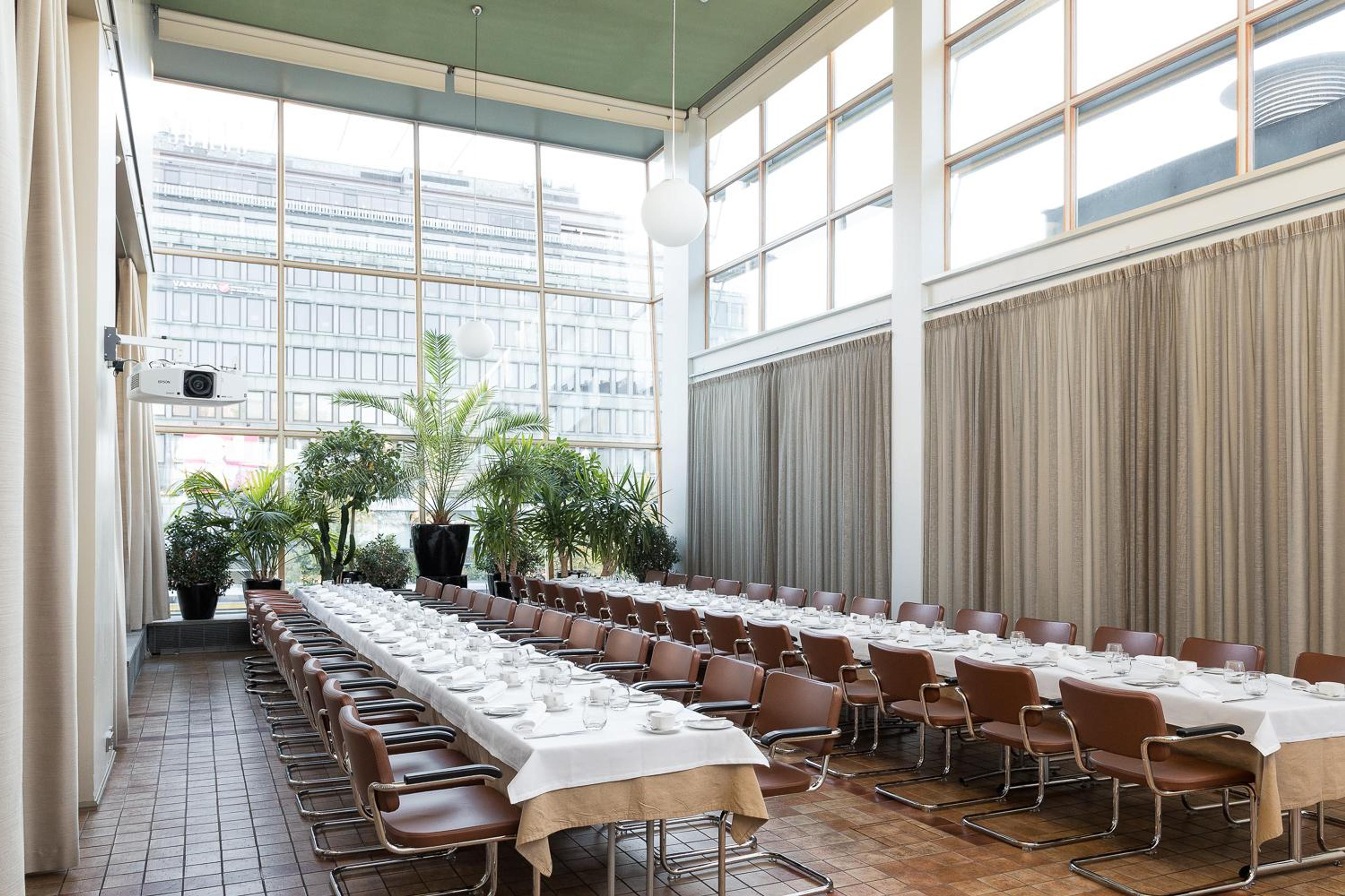 Valoisa kokoustila, jossa pitkä kattaus, ruskeat tuolit, suuret ikkunat ja viherkasveja. Tilassa on tiilivaaleat lattialaatat ja videotykki.