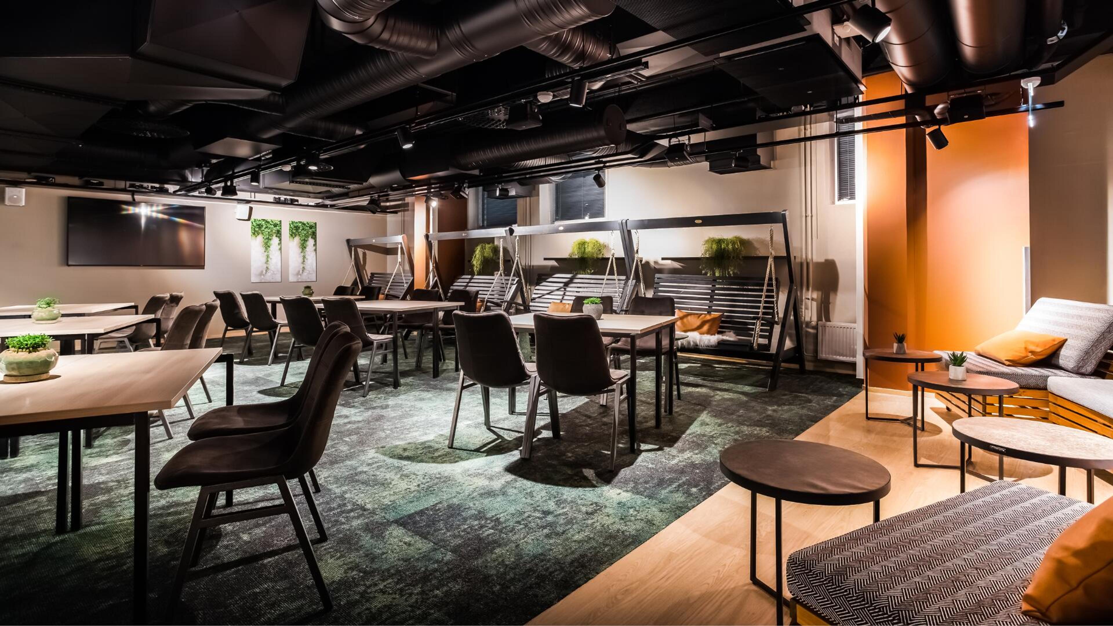 Moderni kokoustila, jossa pöytiä ja tuoleja, mukavia keinuvia sohvaa, viherkasvi-elementtejä ja näyttö seinällä. Tasainen lattiapinta.