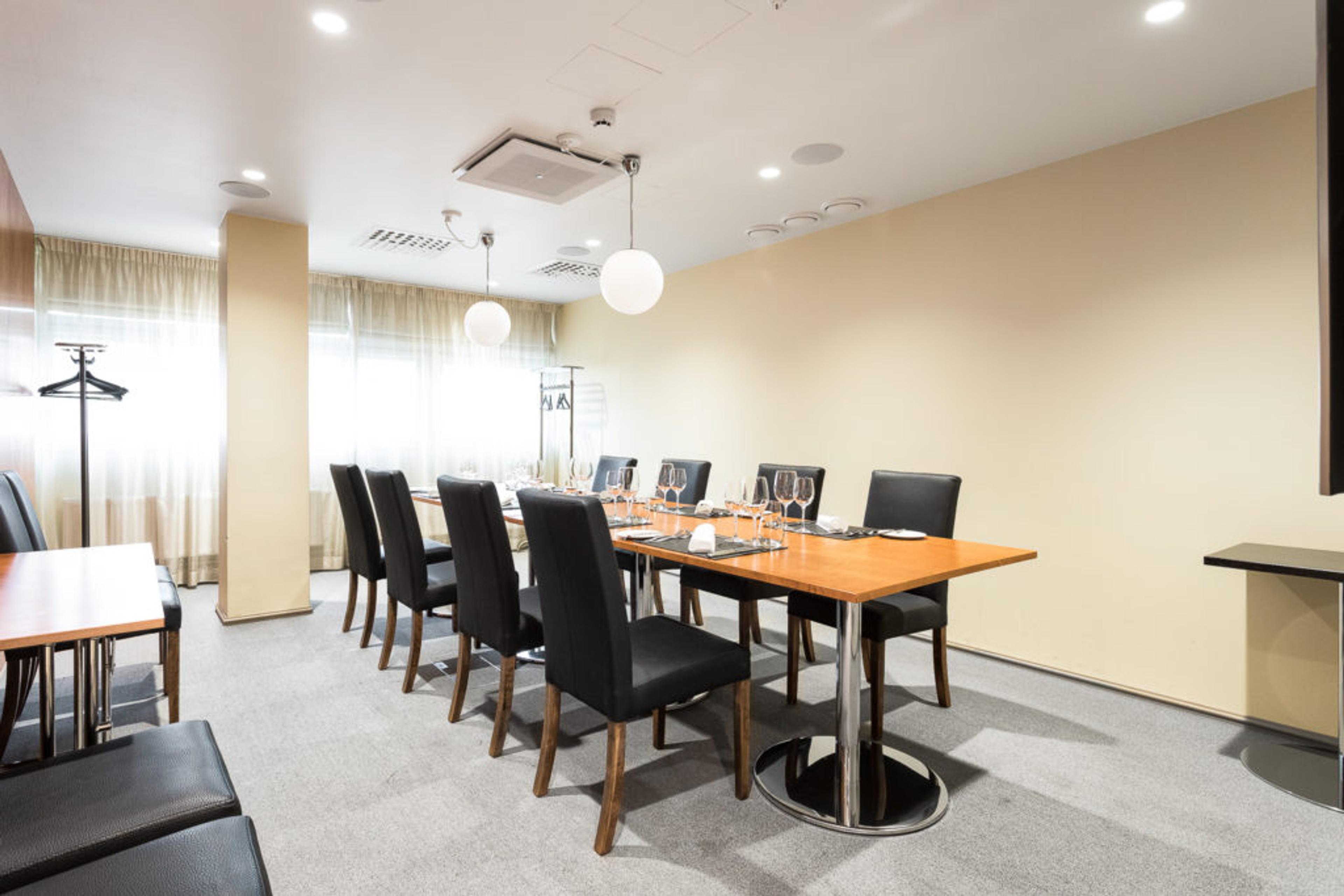 Valoisa kokoushuone, jossa pitkä puinen pöytä, mustat tuolit, viinilasit ja tarjoilu. Huoneessa vaaleat seinät ja laskeutuvat valaisimet.