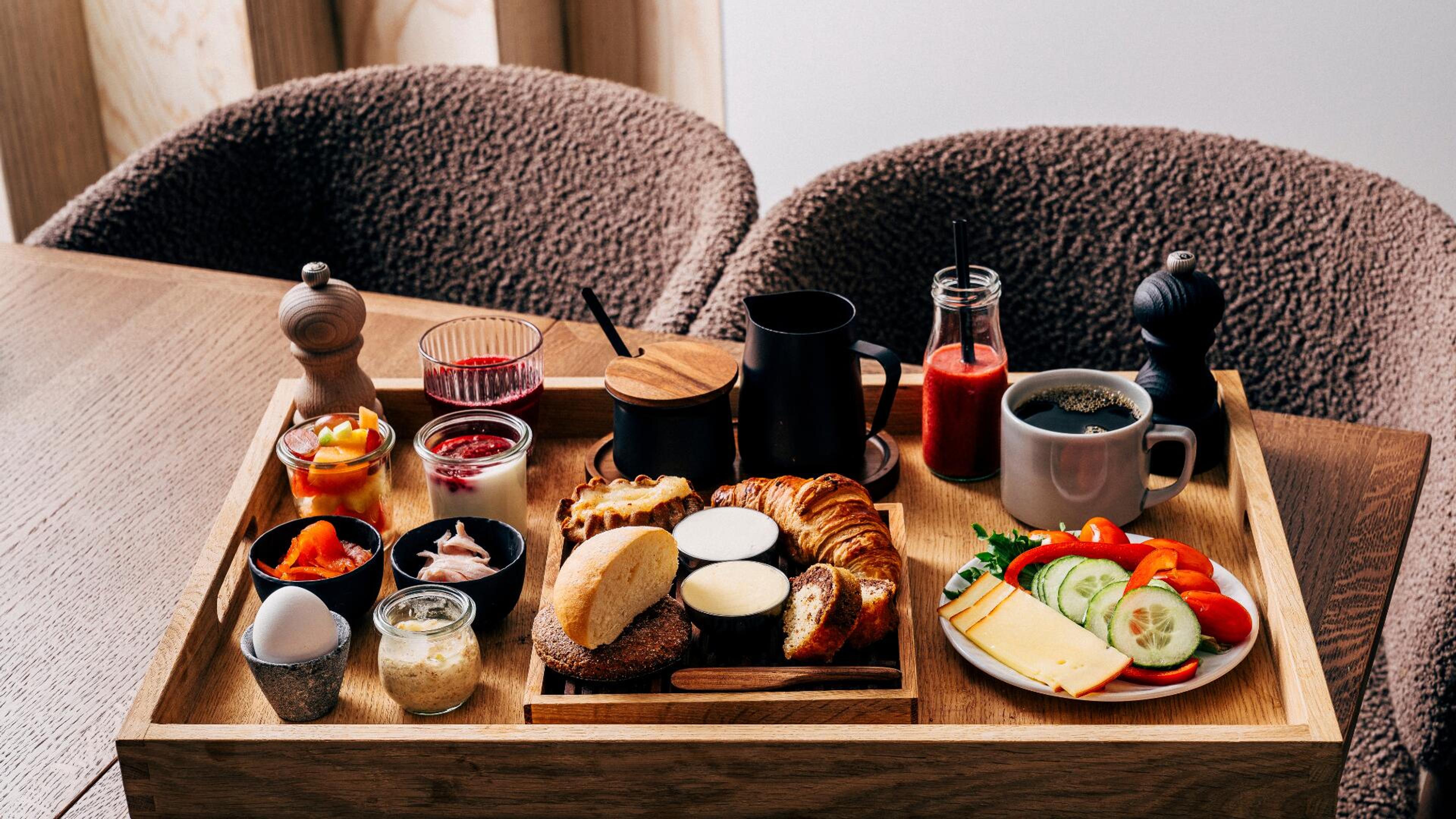 Runsas aamiainen puisella tarjottimella. Tarjolla leipää, croissant, kahvia, mehua, jogurttia, munia, juustoa, vihanneksia ja hedelmiä.