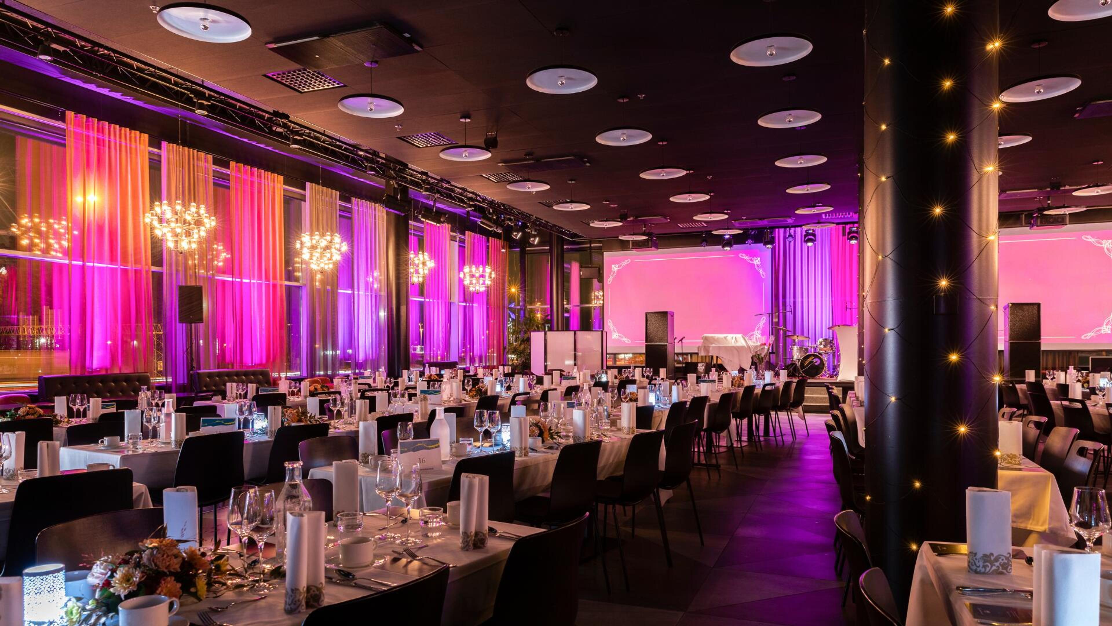 Juhlasali pinkillä valaistuksella, katossa pyöreät valaisimet. Pitkillä pöydillä valkoisia pöytäliinoja ja koristeluita.