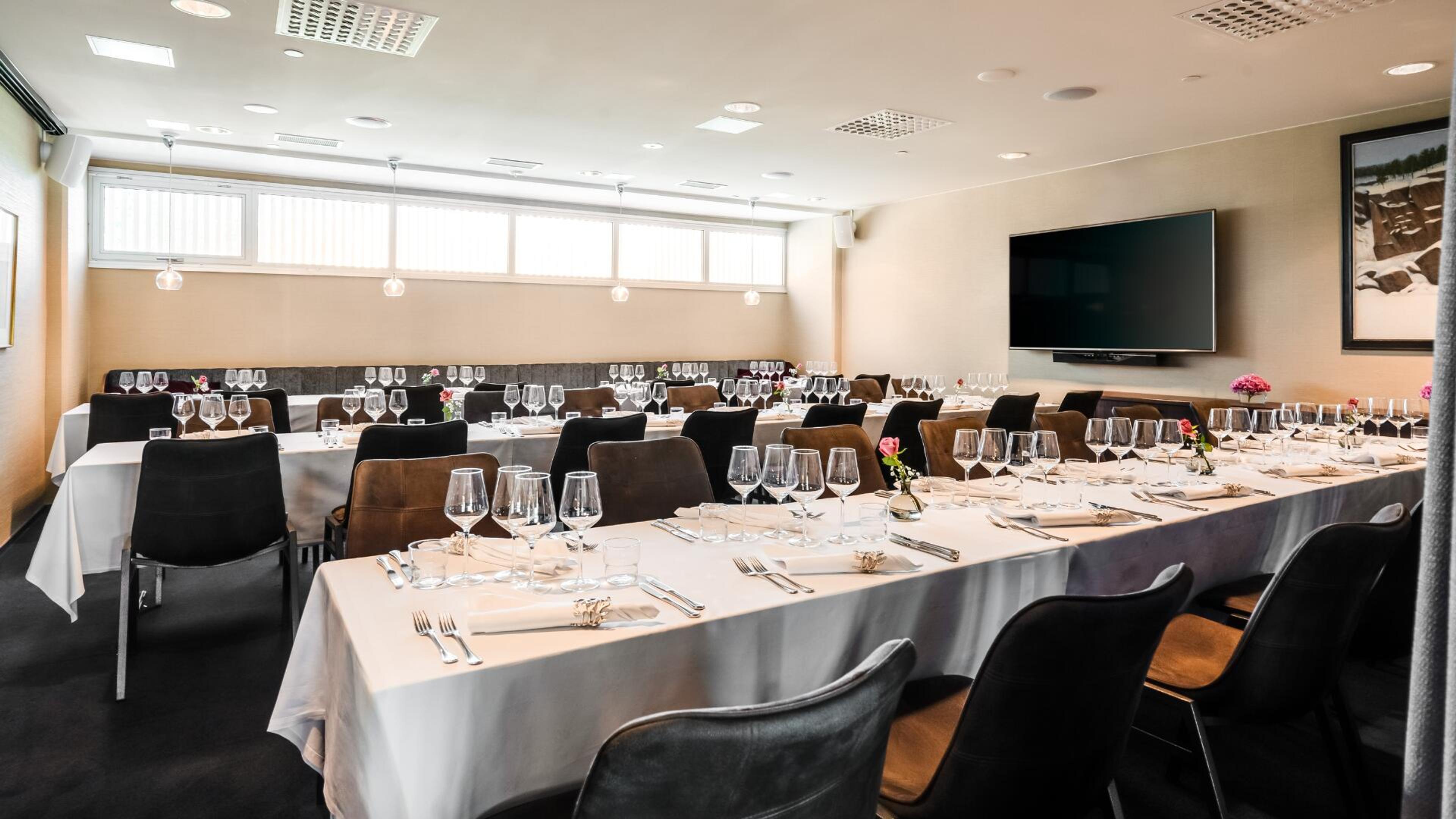 Tyylikäs yksityinen ruokailutila valkoisin pöytäliinoin ja kattauksin. Mustia ja ruskeita tuoleja, seinällä televisio ja tauluja.