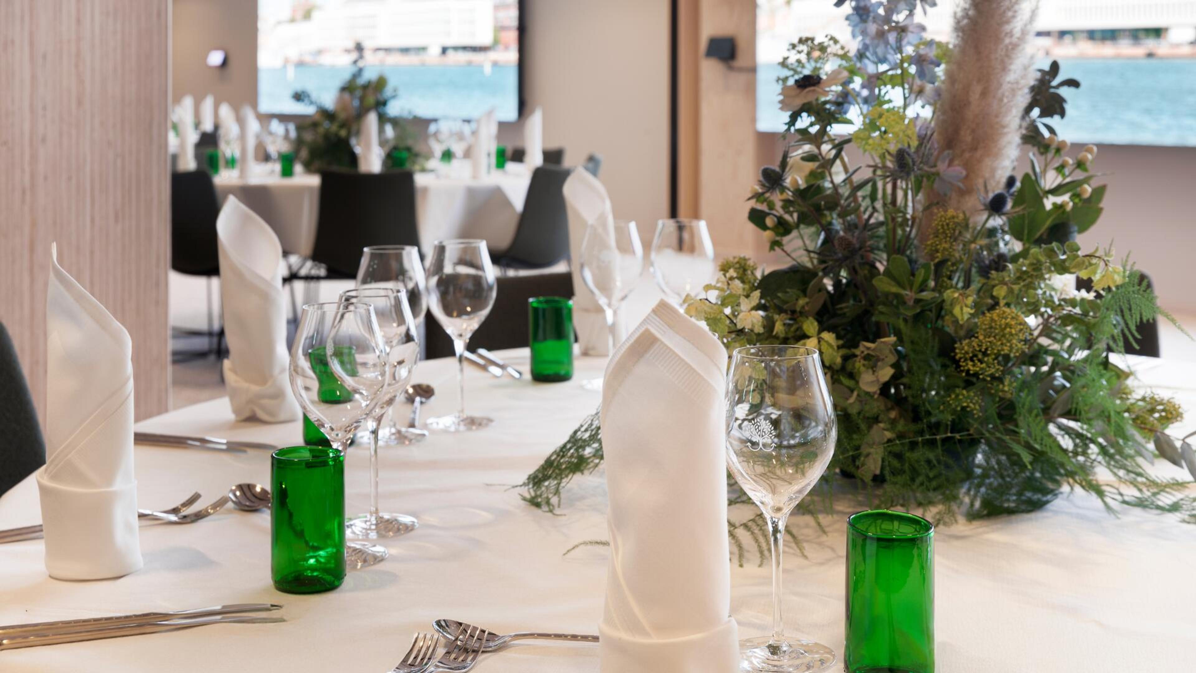 Tyylikkäästi katettu ravintolan pöytä valkoisella liinalla, vihreitä pulloja, viinilaseja ja luonnonkukka-asetelma. Taustalla merinäkymä ikkunoista.