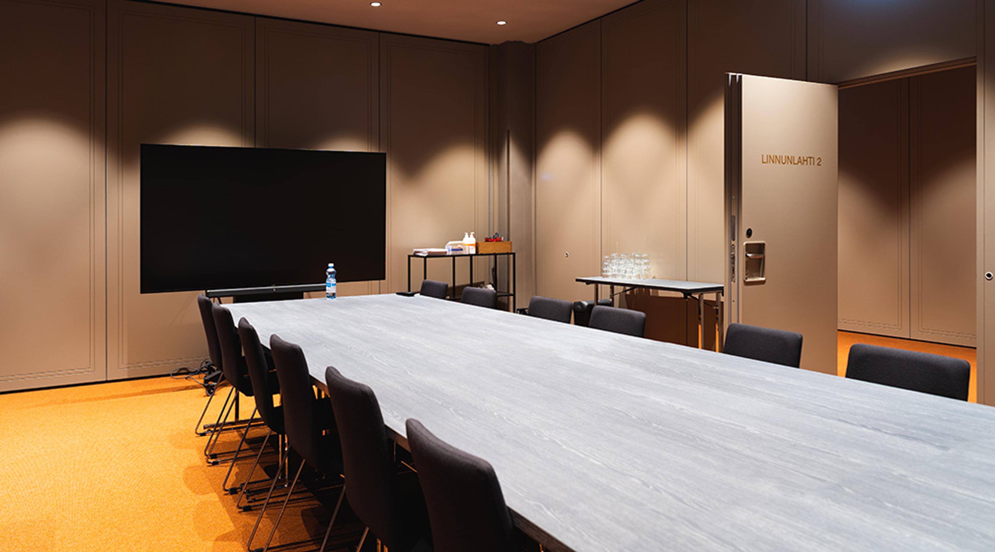 Modern meeting room with beige walls, long light-colored table and black chairs. Large screen in corner and floor covered with orange carpet.