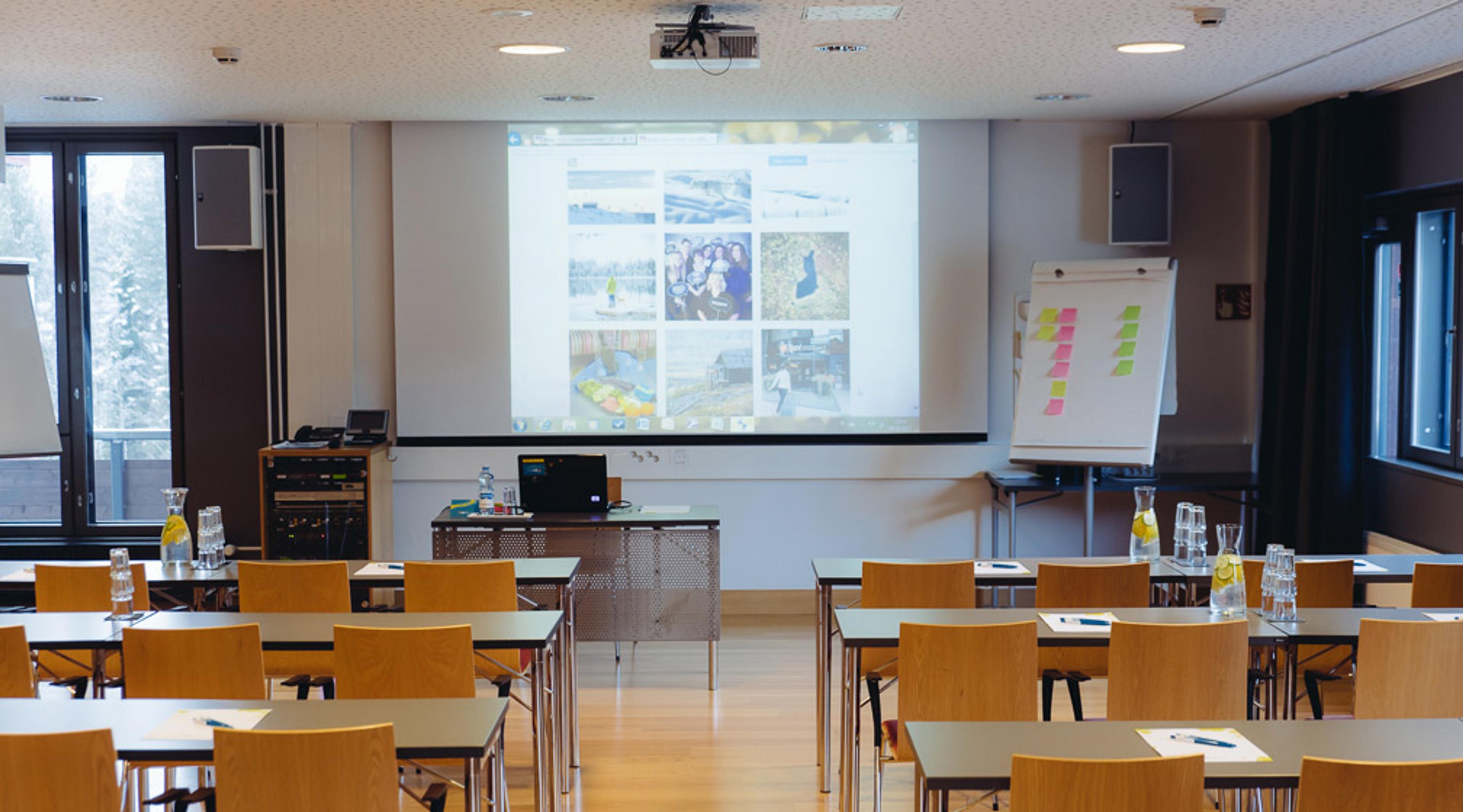 Training room with wooden chairs, projector and whiteboard at front, winter scenery visible through windows. Wooden floor, drinks and papers on tables.