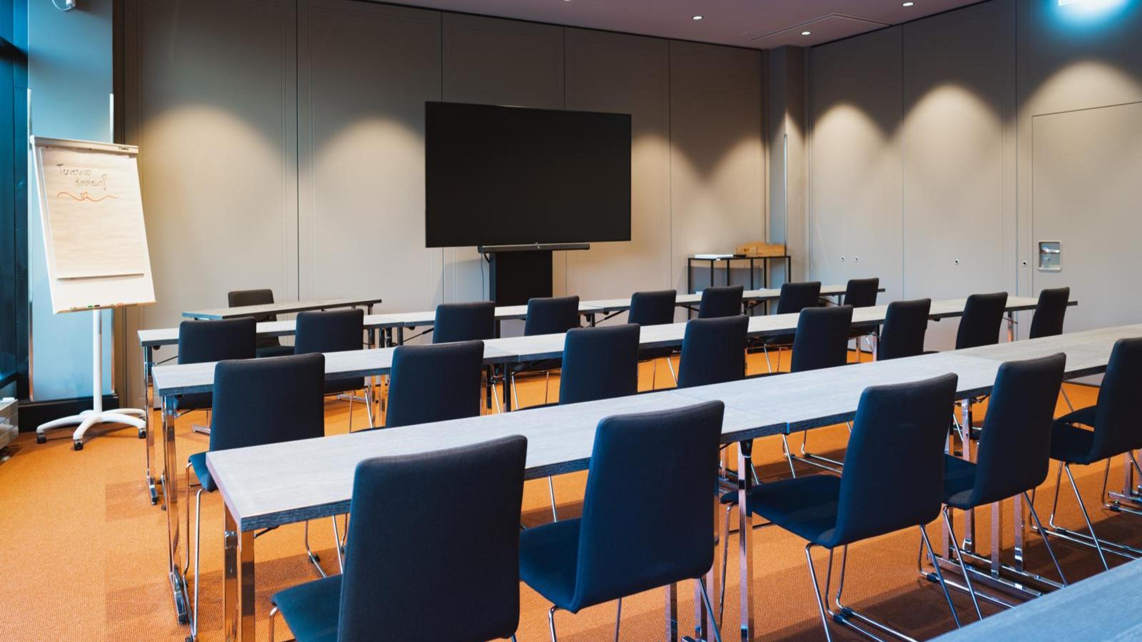 Conference room with rows of tables and blue chairs, large display screen at front and a flipchart to the left. Orange wall-to-wall carpet.