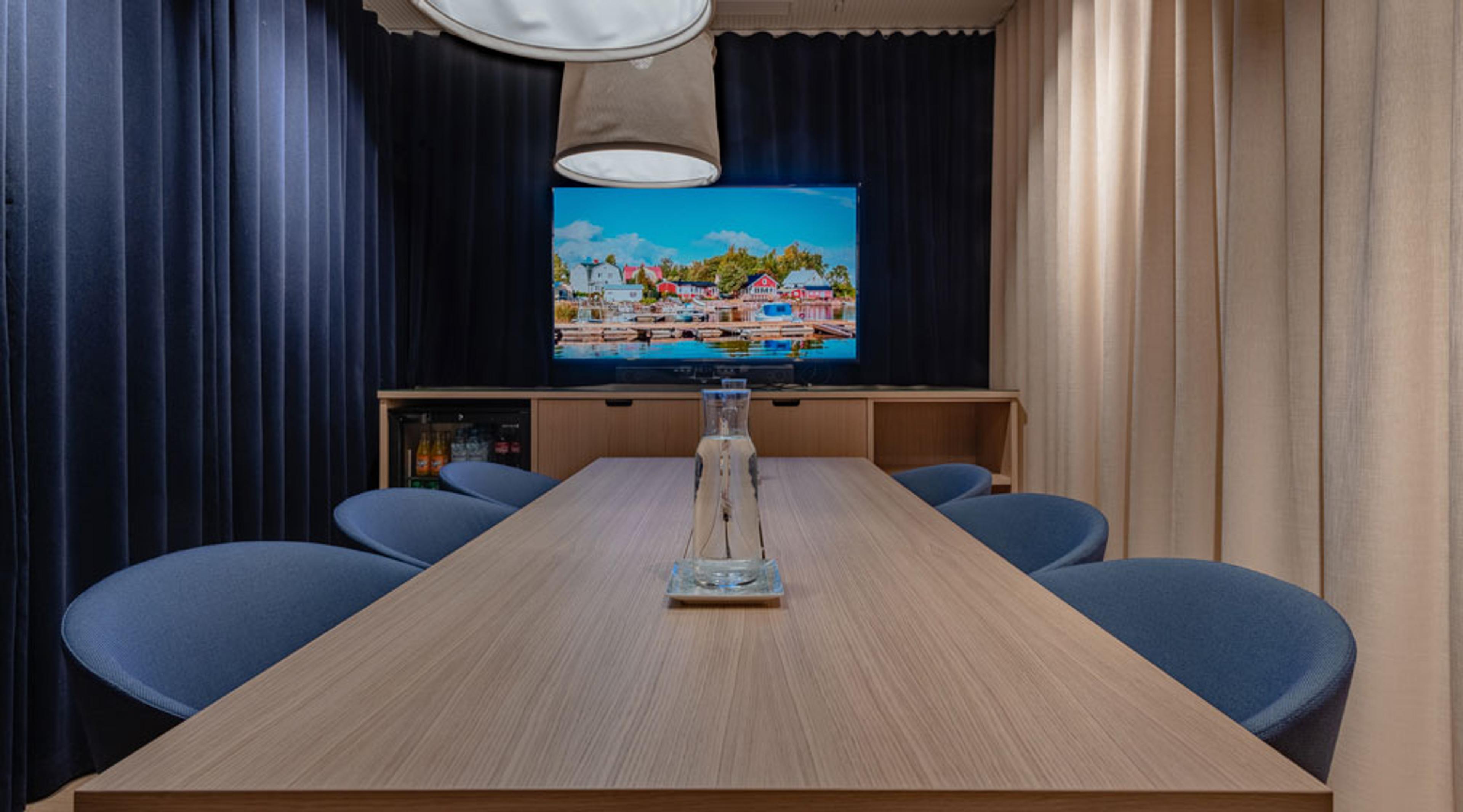 Moderni neuvotteluhuone puisella pöydällä, sinisillä tuoleilla ja television edessä. Vaaleat ja siniset verhot ympäröivät tilaa.