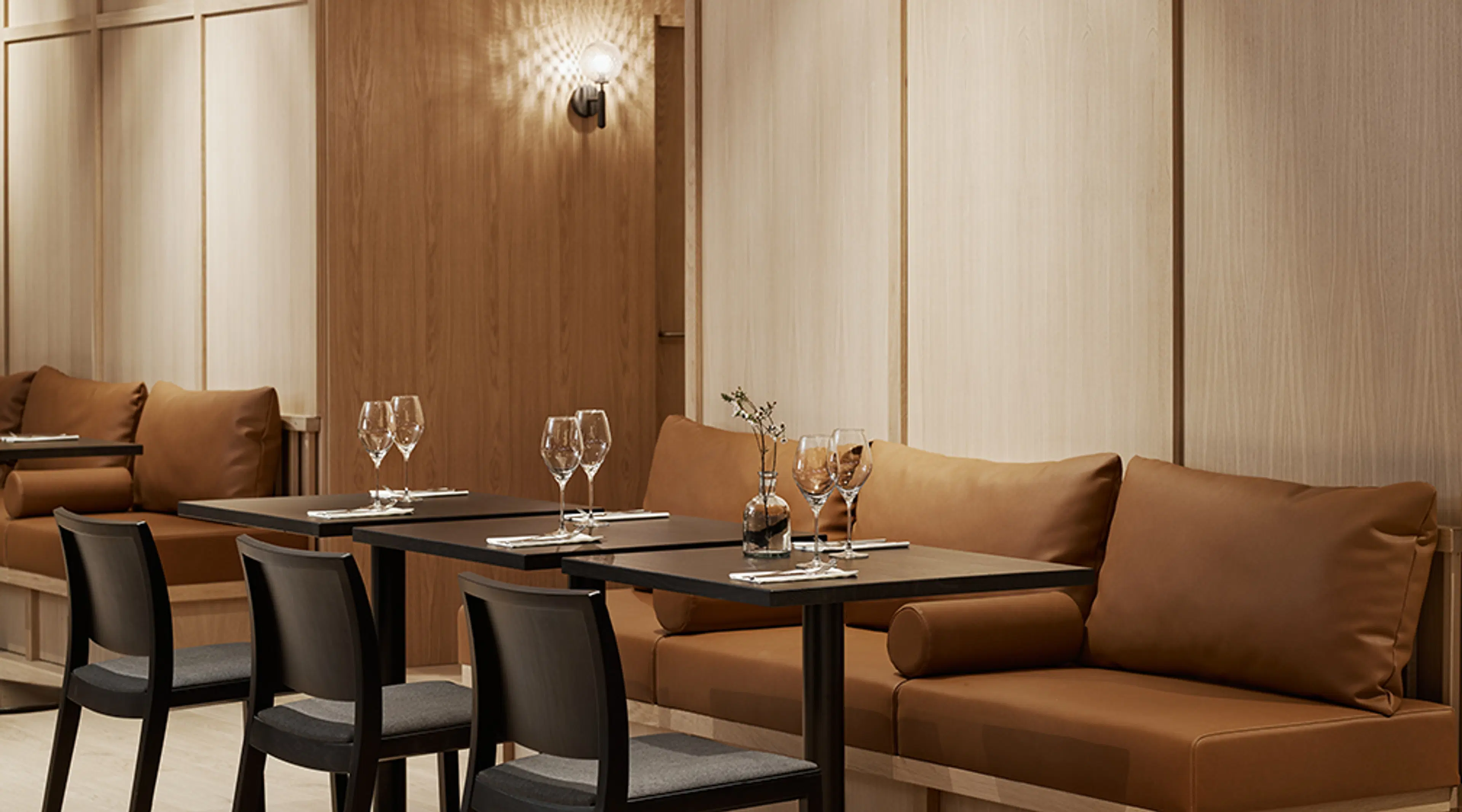 Moderni ravintola sisustettuna puupaneloinnilla, ruskeilla sohvilla ja mustilla pöydillä. Pöydillä viinilaseja ja kattauksia.