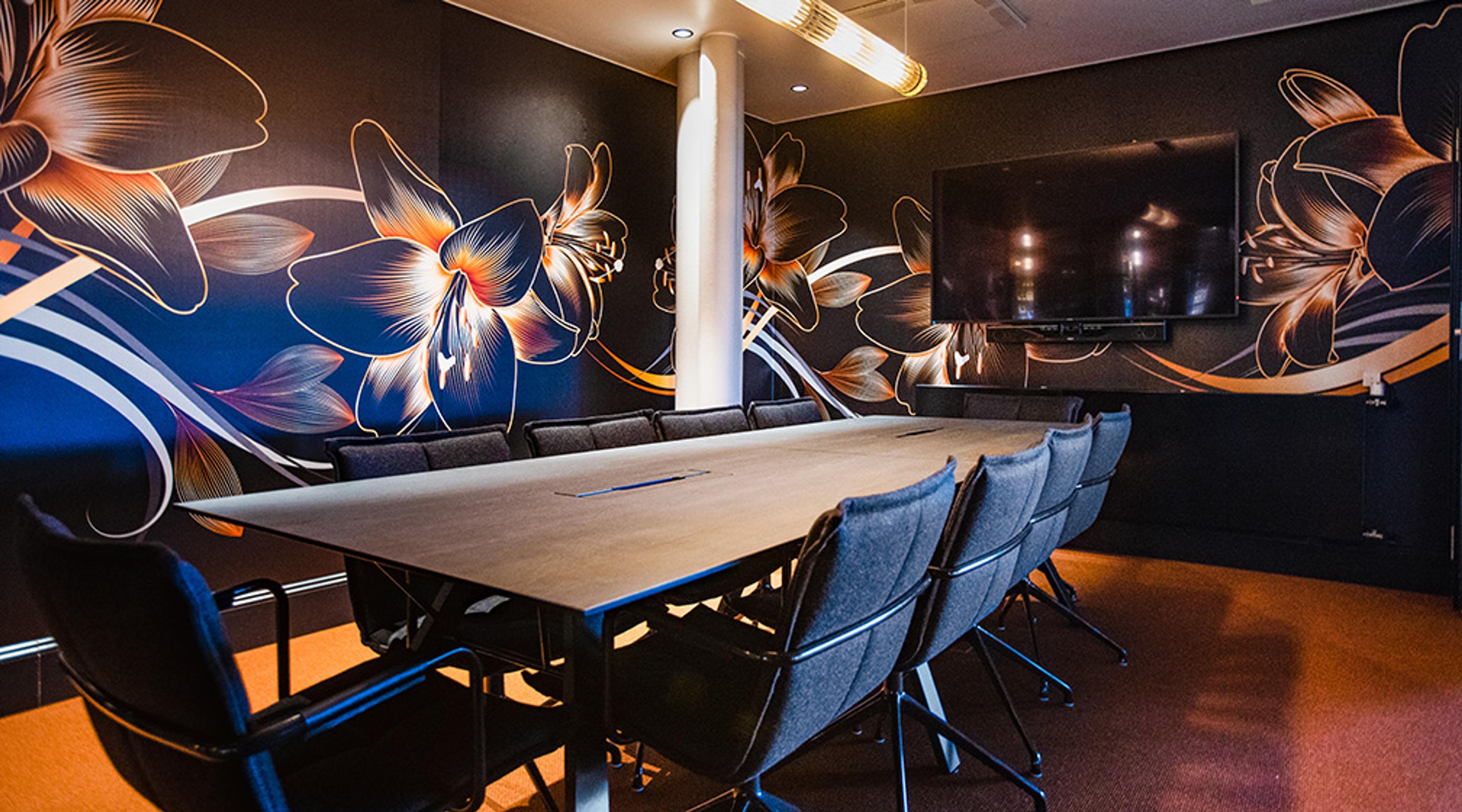 Meeting room with wooden table, grey chairs, large floral murals on dark walls and a TV screen. Even floor and good lighting.