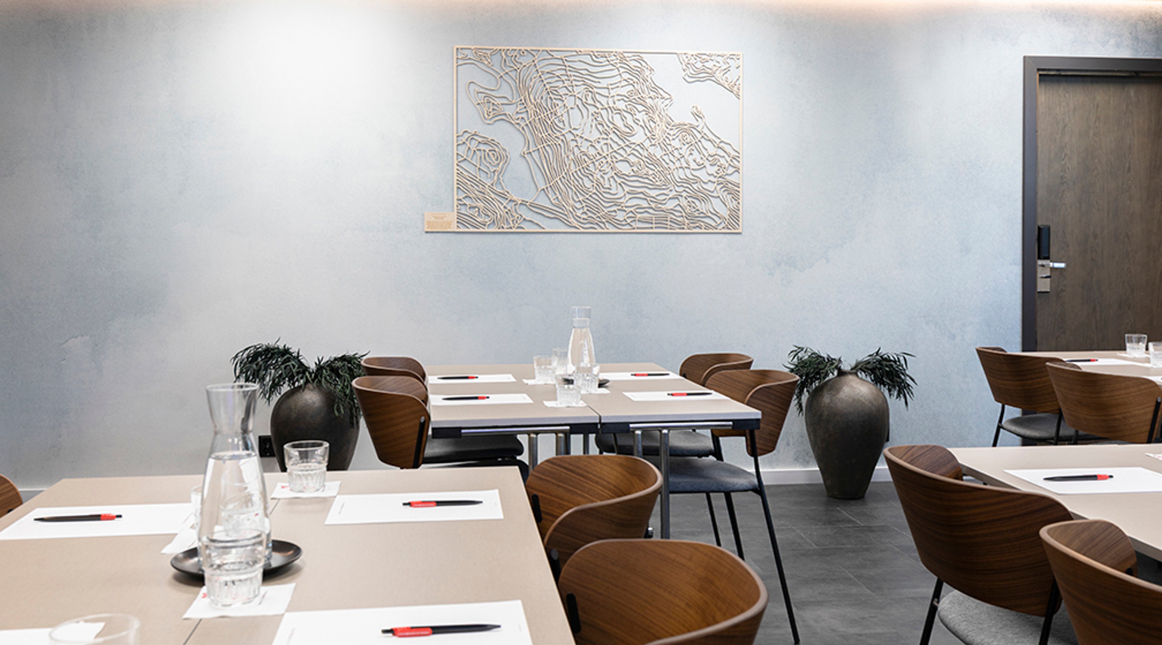 Moderni kokoustila vaaleansinisellä seinällä, jossa taideteosta. Puiset tuolit ja pöydät valmiina kokoukseen, vesikarahveja ja kynät pöydillä.