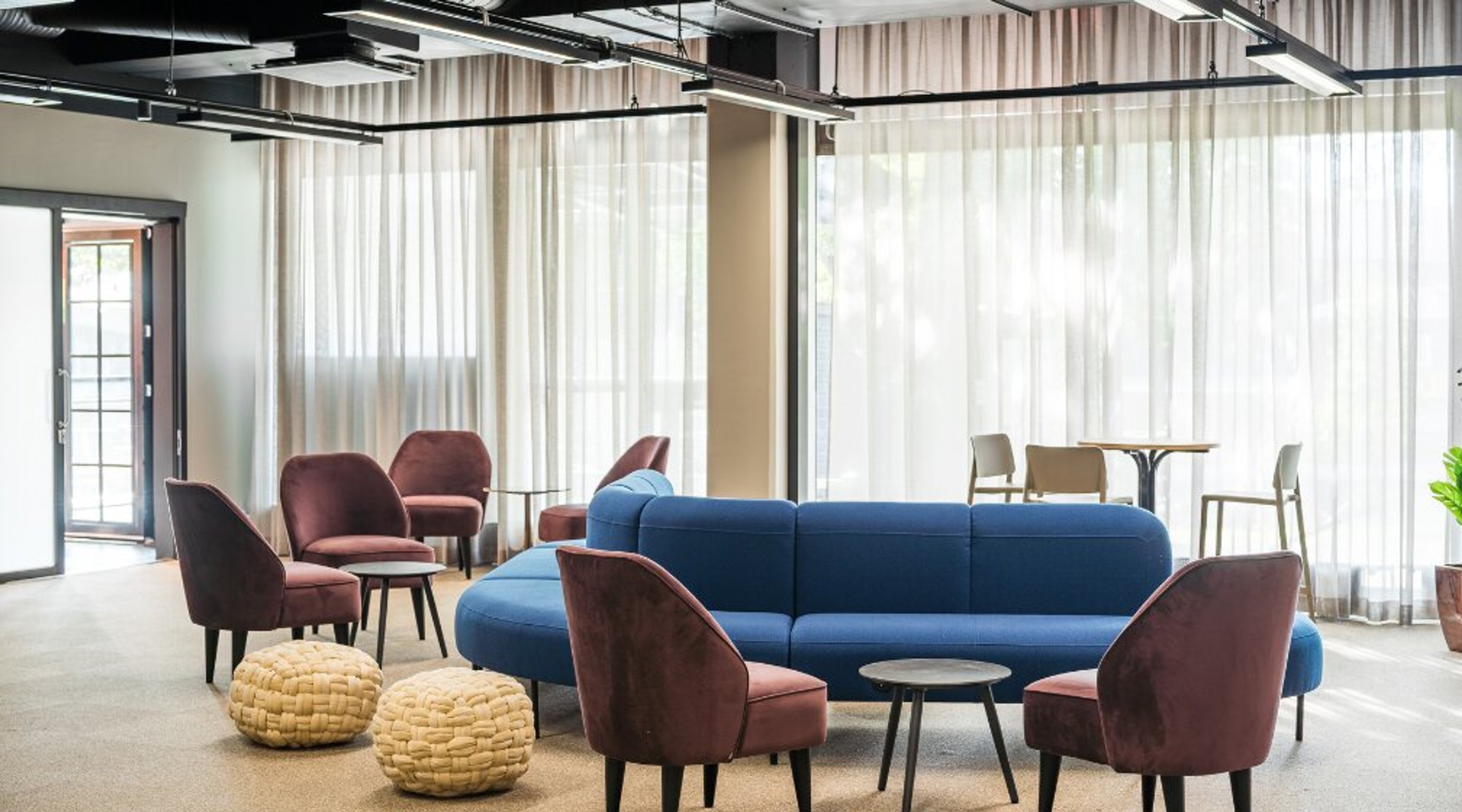 Valoisa lounge-tila, jossa on sininen sohva, viininpunaisia nojatuoleja ja beige matolla. Suuret ikkunaverhot ja tasainen lattiapinta.