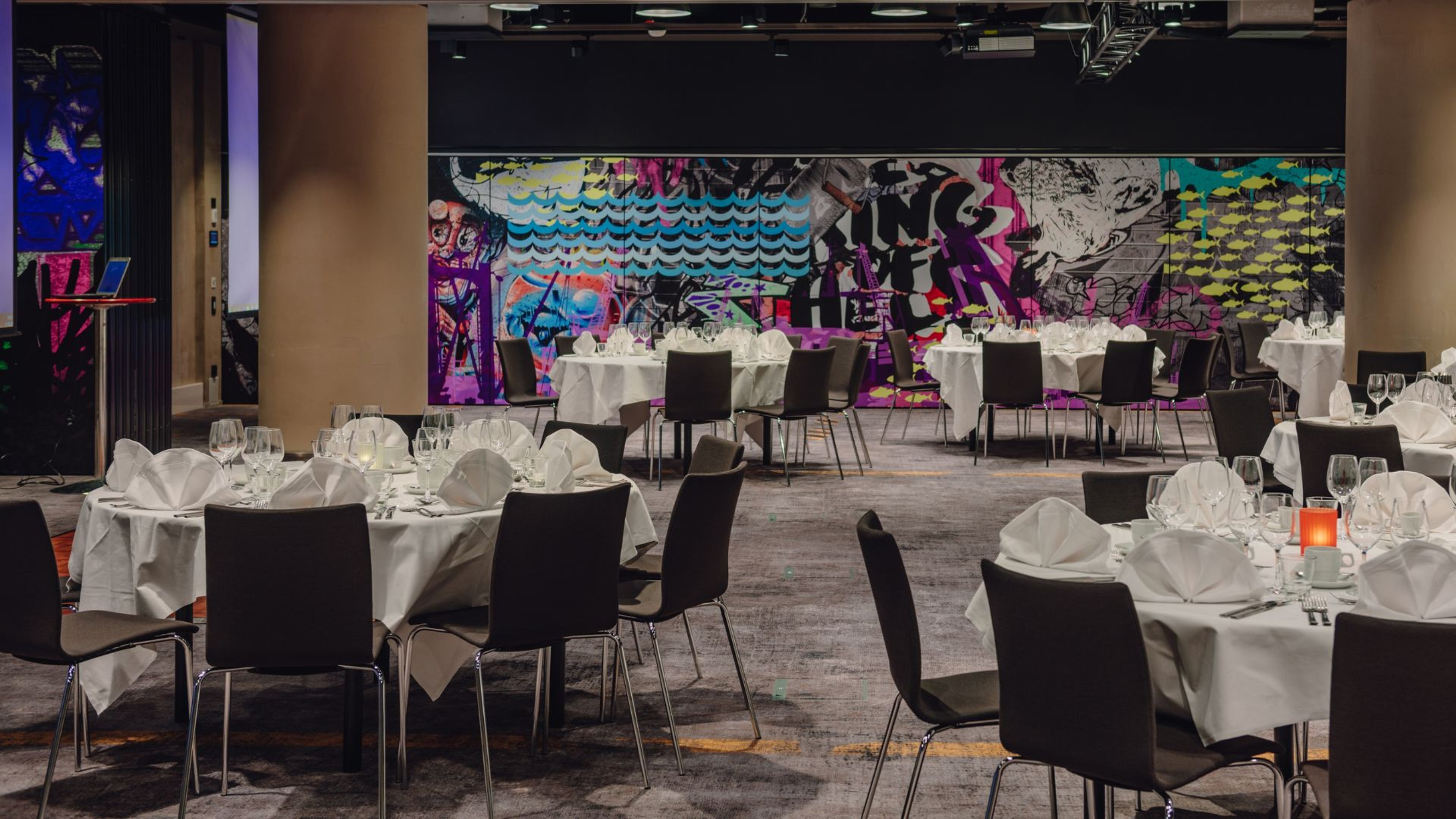 Ravintolasali, jossa on valkoisia pöytiä ja mustia tuoleja. Värikkäät graffitityyppiset seinämaalaukset taustalla. Tasainen lattia ja hyvä valaistus.