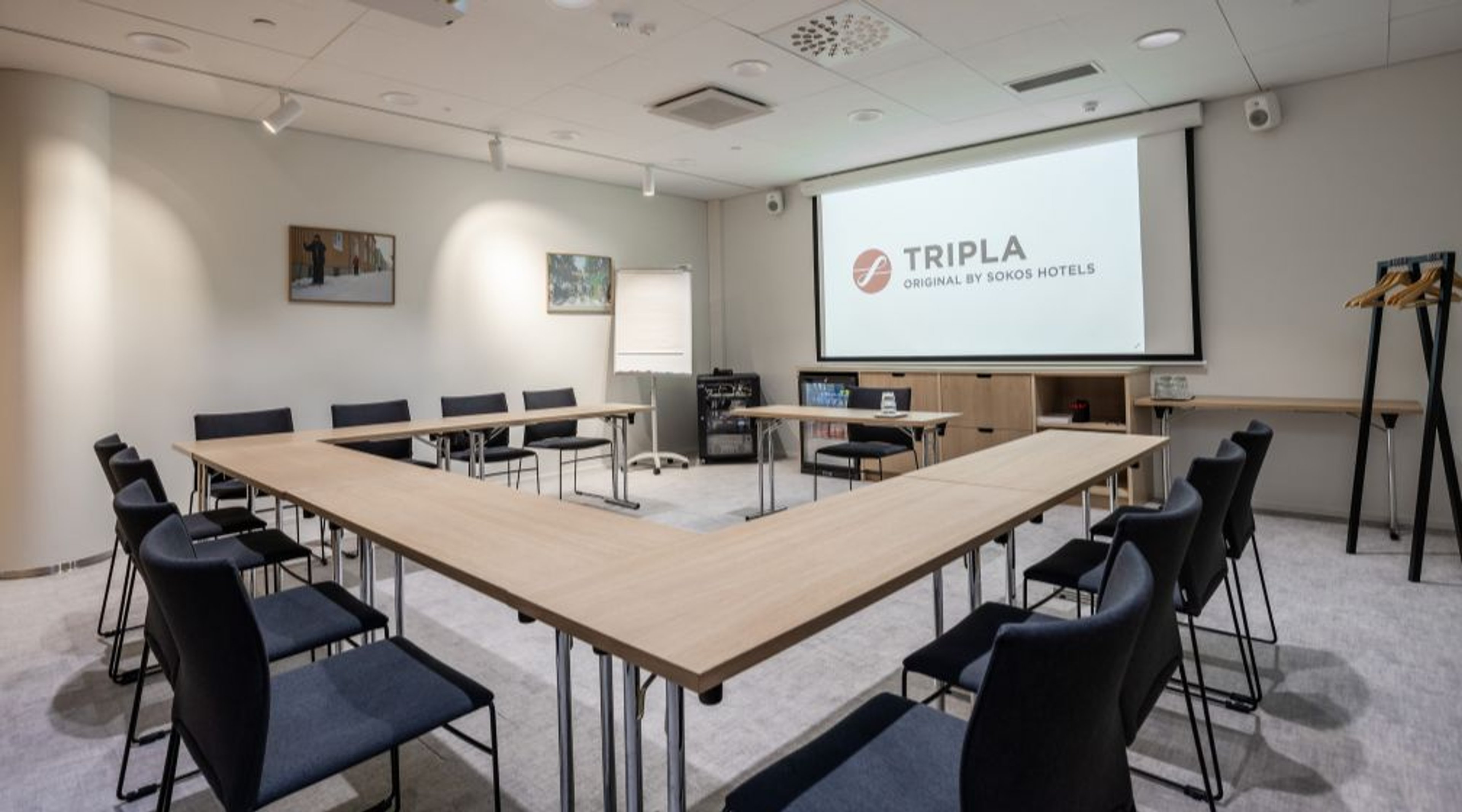 Kokoustila U-muotoisella pöytäasetelmalla, mustilla tuoleilla ja valkokankaalla. Tilassa on hyvä valaistus ja tasainen lattiapinta.