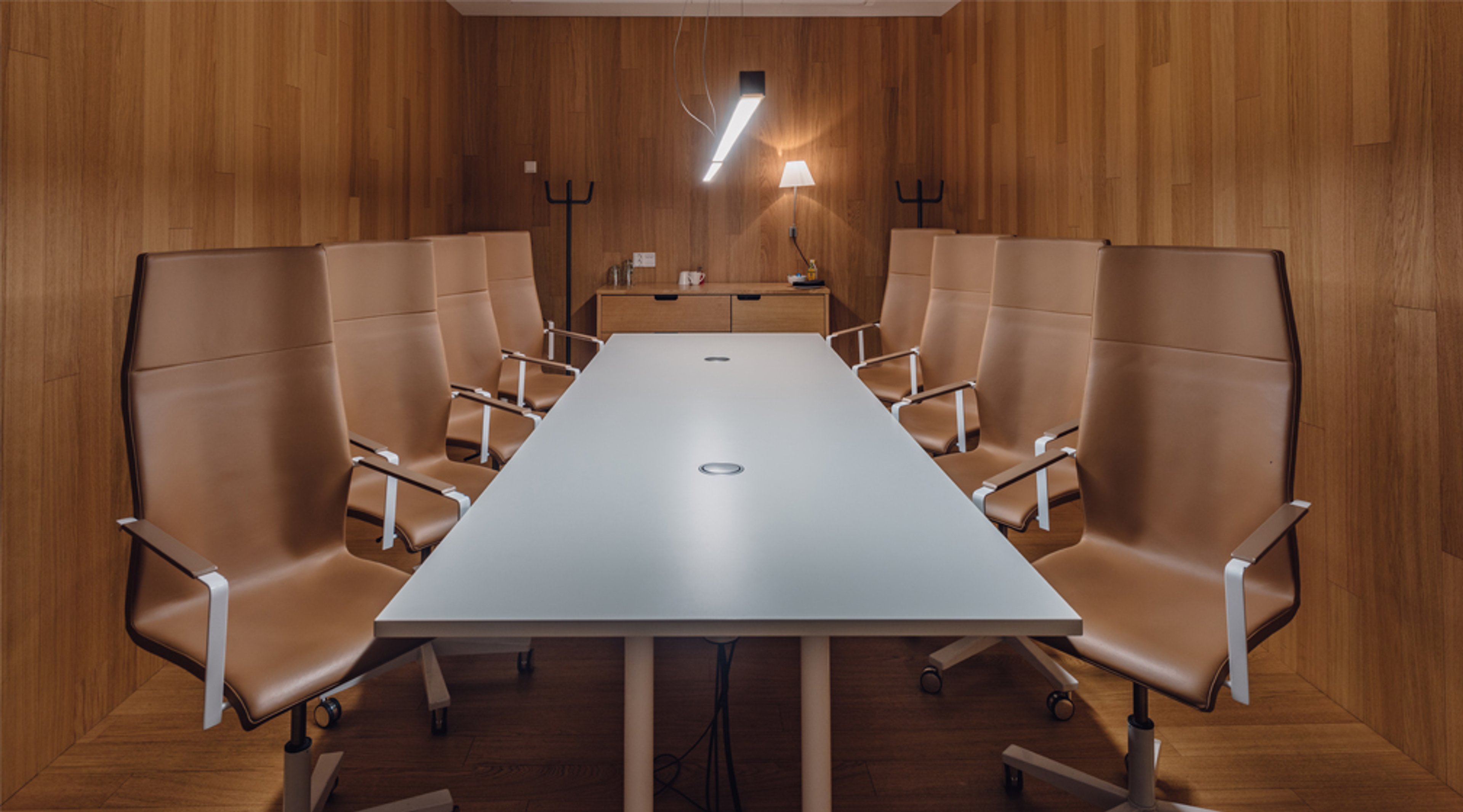 Neuvotteluhuone puisilla seinäpaneeleilla, valkoinen pöytä ja ruskeat tuolit. Lattia puuta, seinällä valaisin ja taustalla sivupöytä.