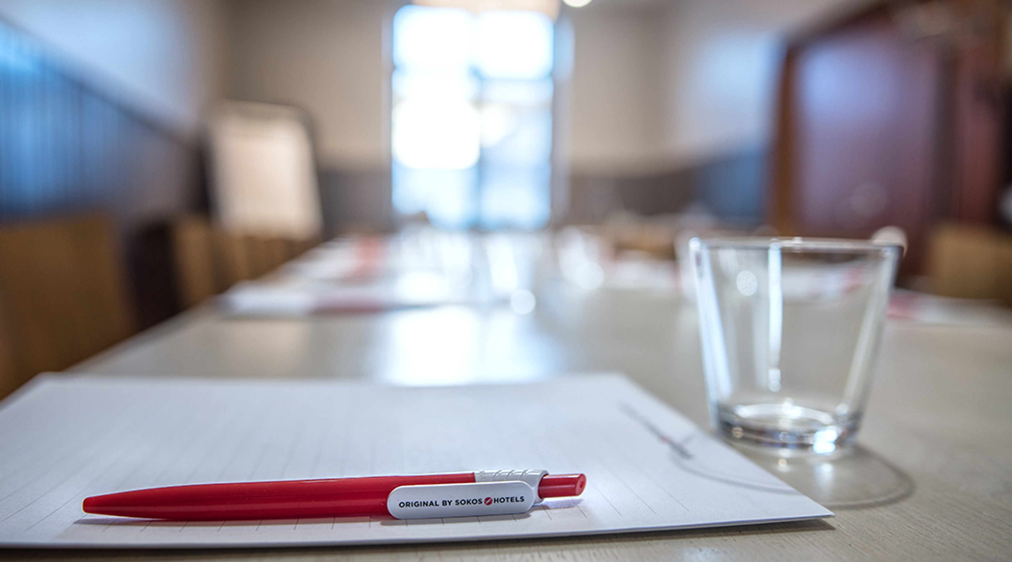 Neuvottelupöytä, jossa punainen kynä ja tyhjä muistilehtiö sekä vesilasi. Taustalla hämärästi näkyy fläppitaulu.