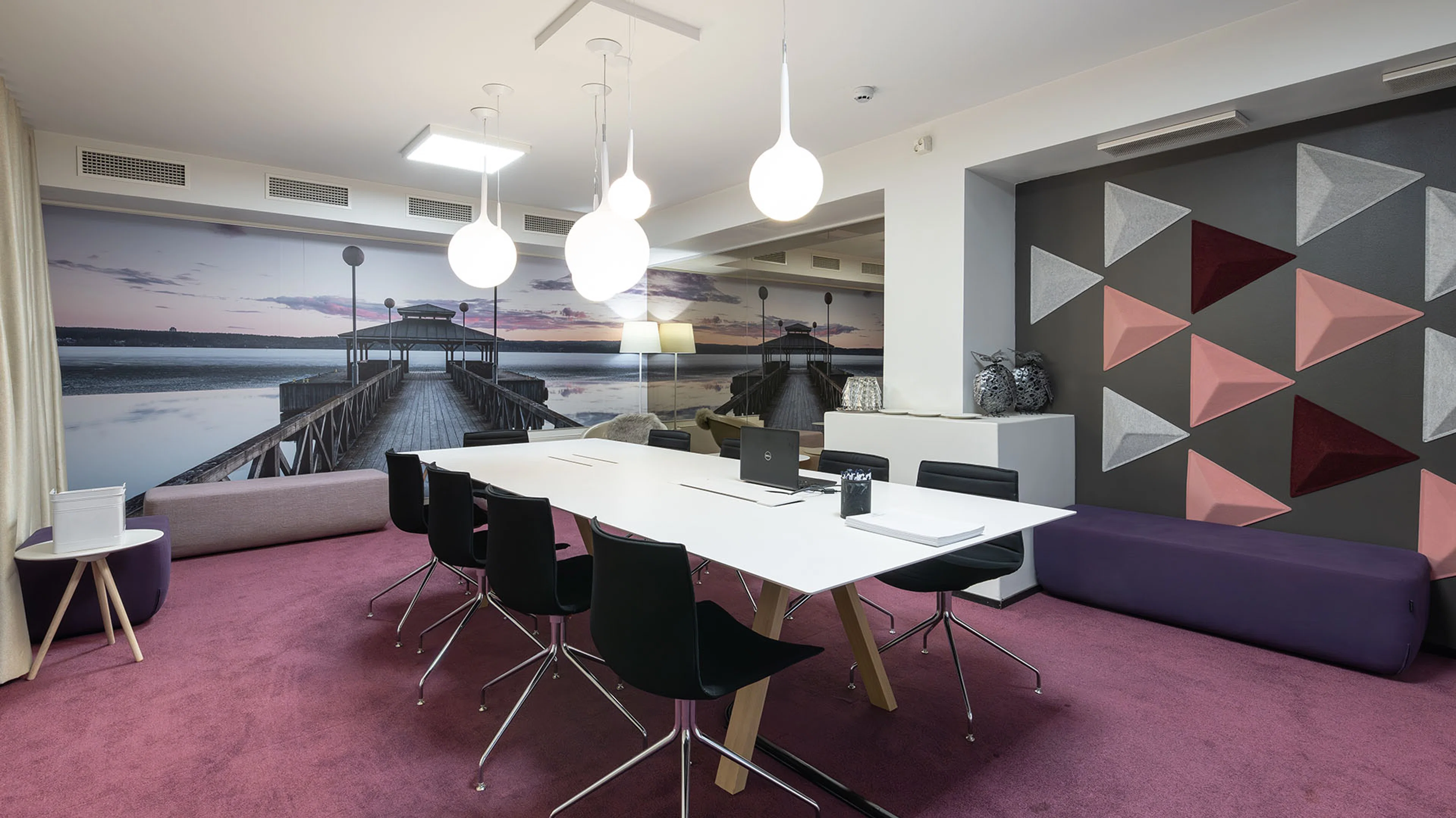 Moderni neuvotteluhuone pinkillä matolla, valkoisella pöydällä ja mustilla tuoleilla. Seinällä laiturista kuvattu järvimaisema ja toisella seinällä geometrinen kuviointi.
