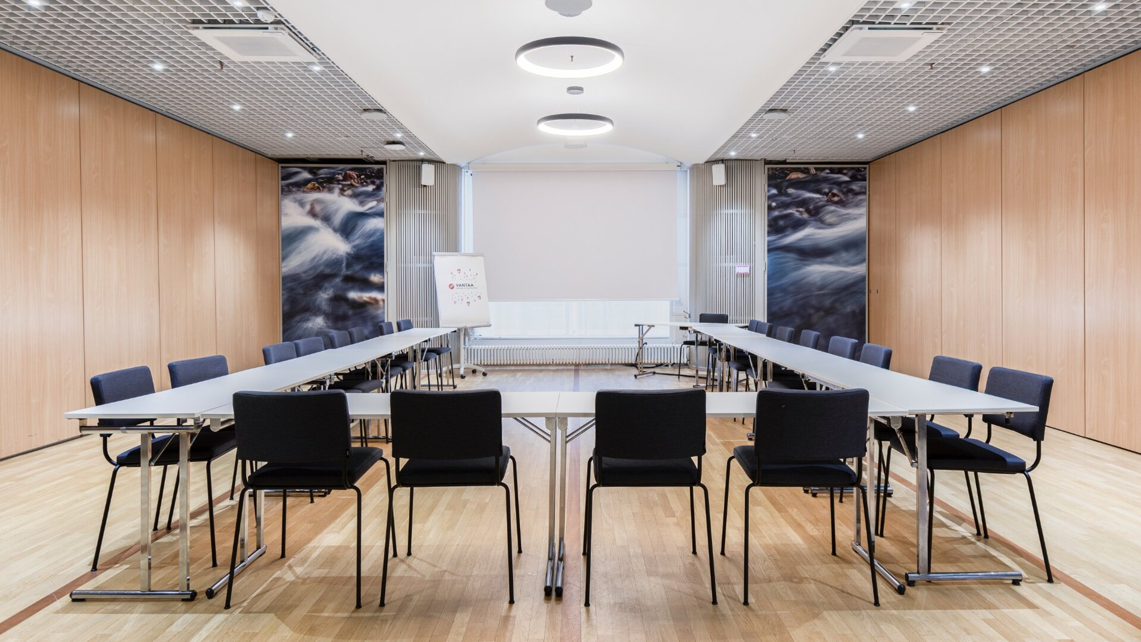 Moderni kokoushuone U-muotoisella pöydällä, mustilla tuoleilla, puisella lattialla ja vesiaiheisilla seinäkuvilla.