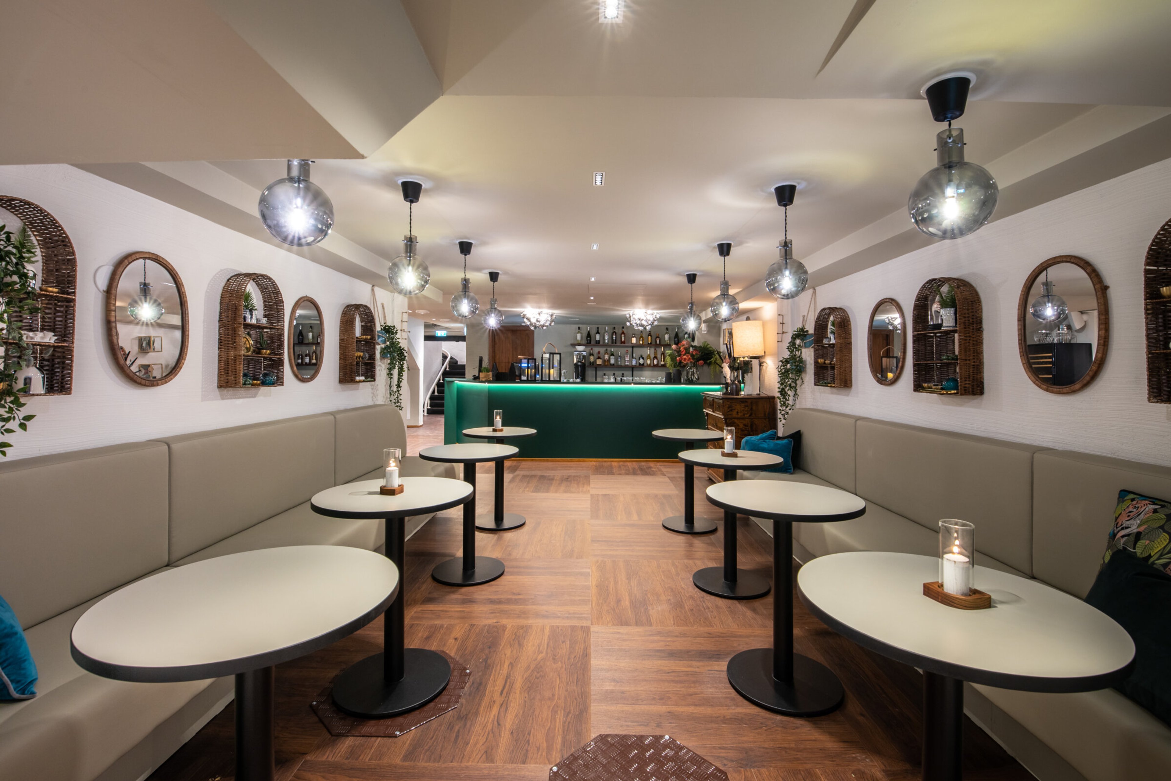 Modern bar with light tables, grey sofas, green bar counter, wooden floor and mirrors with rattan frames on the walls.