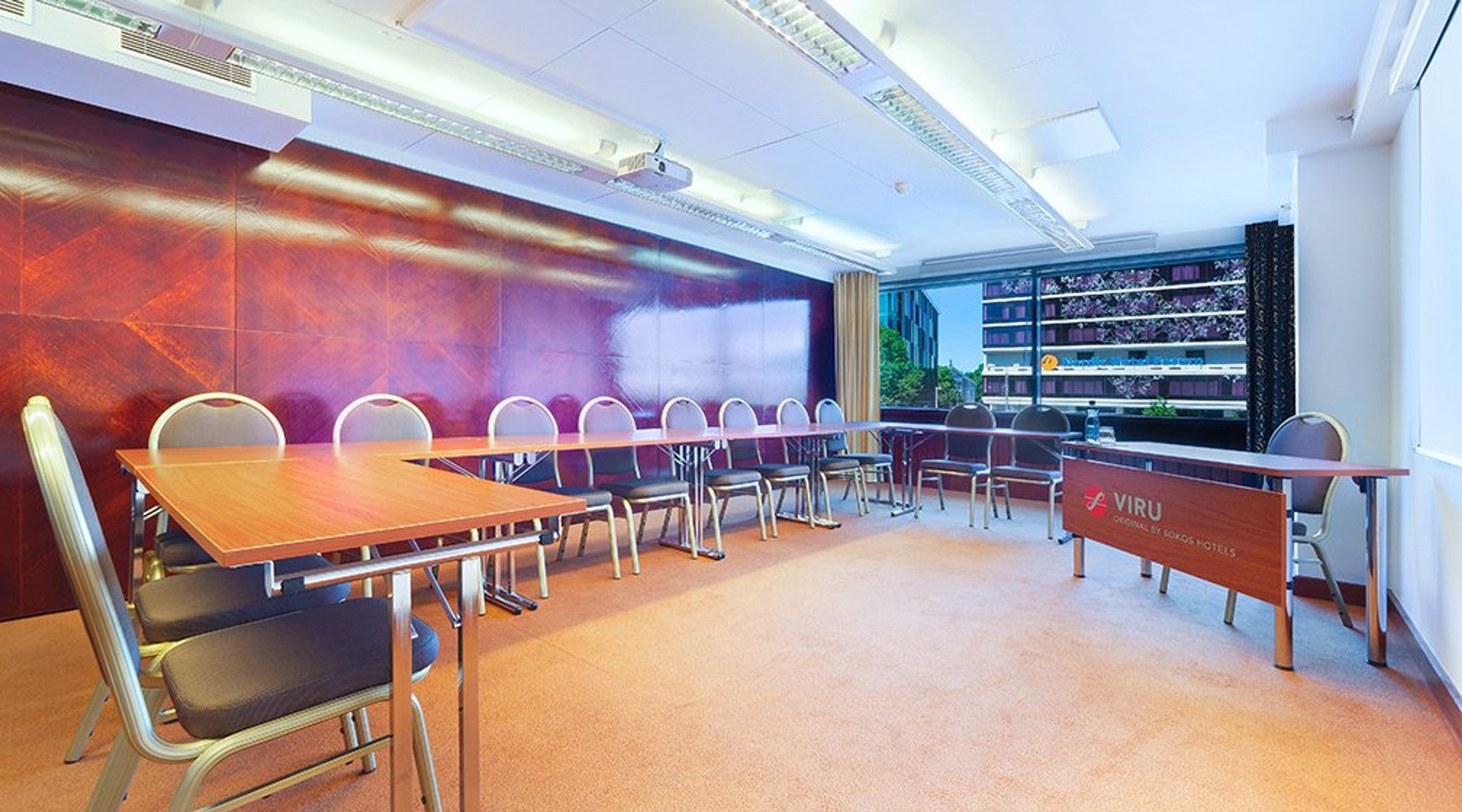 Kokoushuone puisilla pöydillä ja tuoleilla. Punertava seinäpaneeli taustalla, ikkunasta näkyy vihreää kasvillisuutta. Hyvin valaistu tila vaalealla lattialla.