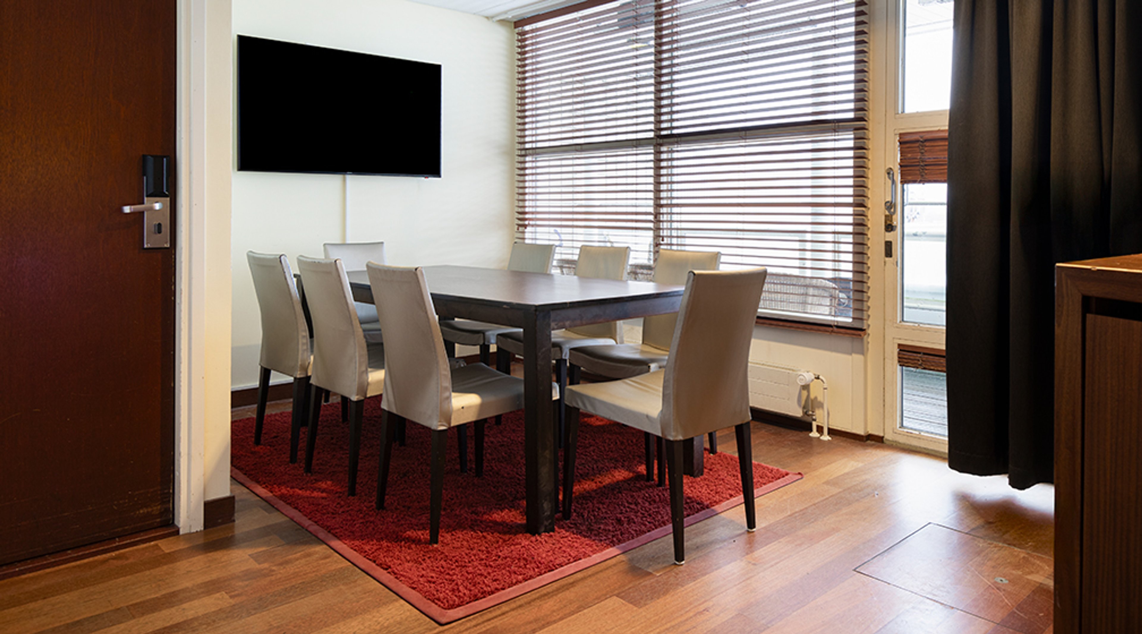Neuvotteluhuone, jossa tumma pöytä ja vaaleat tuolit punaisen maton päällä. Seinällä televisio, ikkunasta tulee luonnonvaloa. Puulattia ja lasiovi.