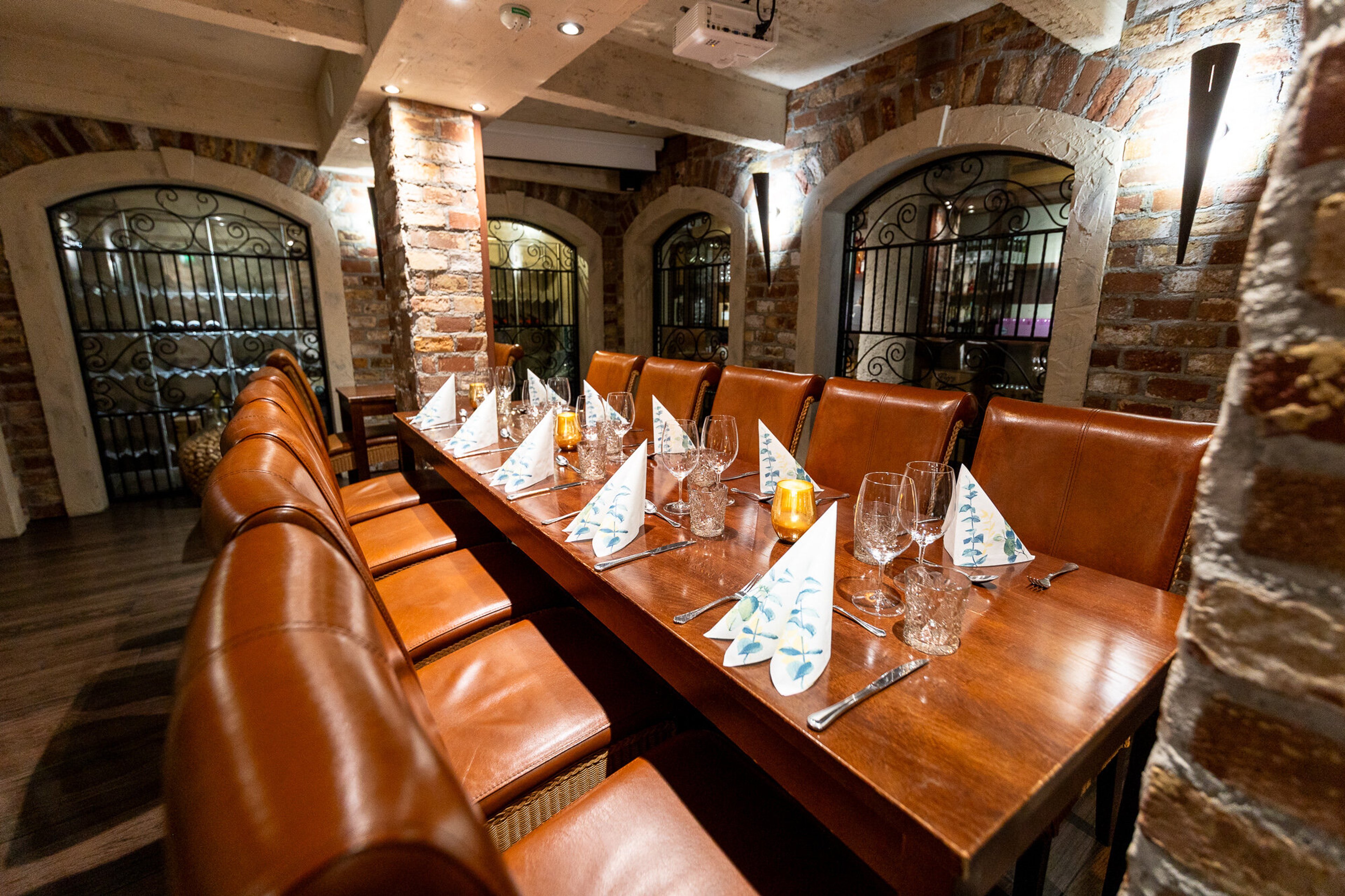 Tunnelmallinen ravintolan yksityishuone tiiliseinillä, pitkä puupöytä kattauksilla ja oransseilla nahkatuoleilla. Taustalla koristeelliset rautaiset ikkunat.