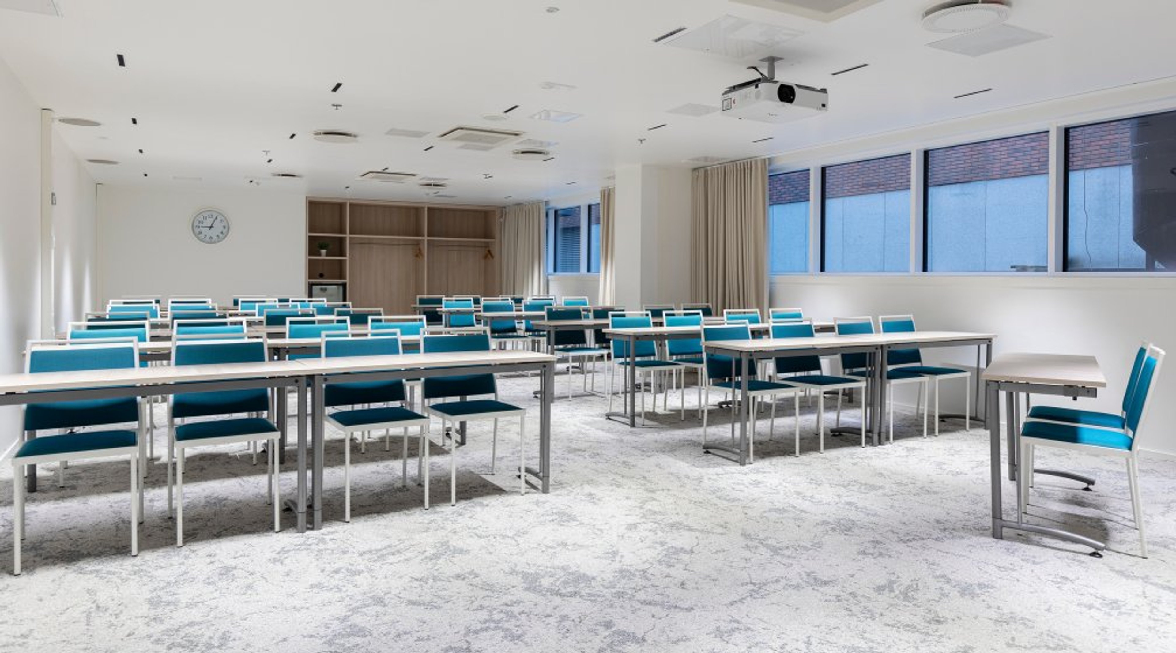 Valoisa kokoustila sinisillä tuoleilla ja valkoisilla pöydillä. Seinällä kello ja kattoon asennettu projektori. Lattialla vaaleanharmaa matto.