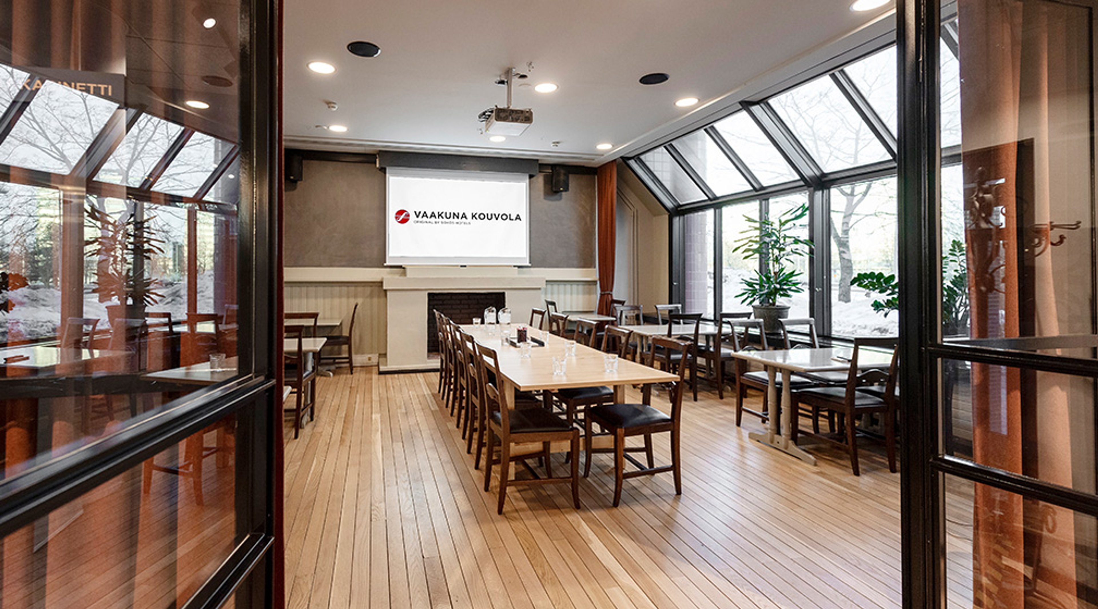 Kokoustila puisella lattialla, pitkä pöytä keskellä ja tuoleja ympärillä. Isot ikkunat tuovat luonnonvaloa, valkokankaan edessä.