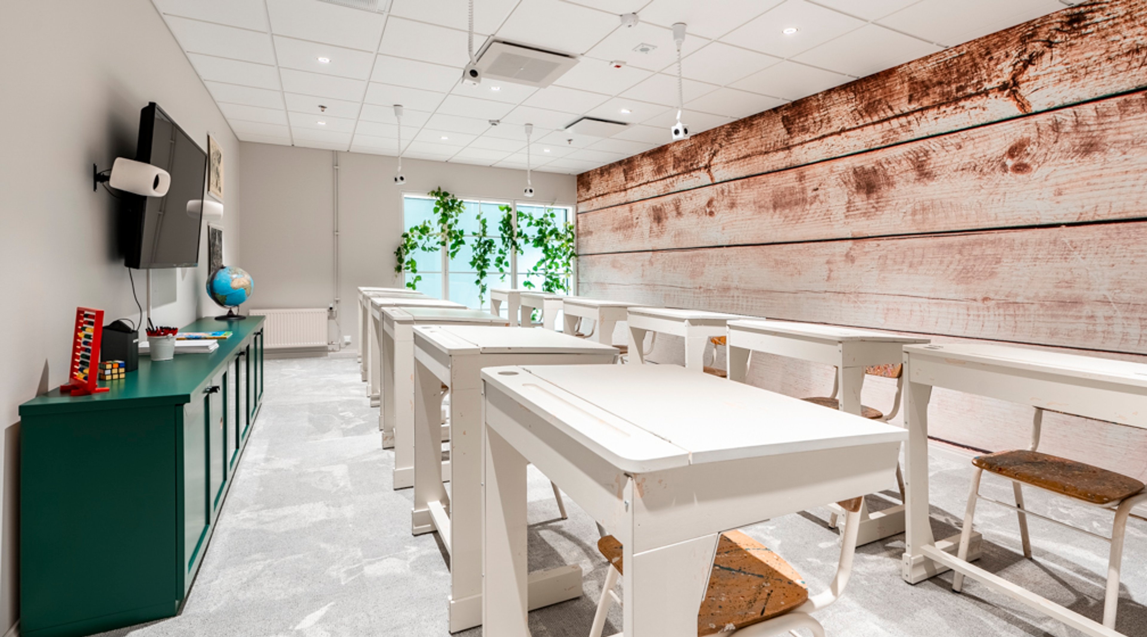 Moderni luokkahuone, jossa valkoiset pöydät, rustiikkinen puuseinä, vihreä säilytyskaluste, television ja vihreää kasvillisuutta ikkunalla. Tasainen lattia ja hyvin valaistu tila.