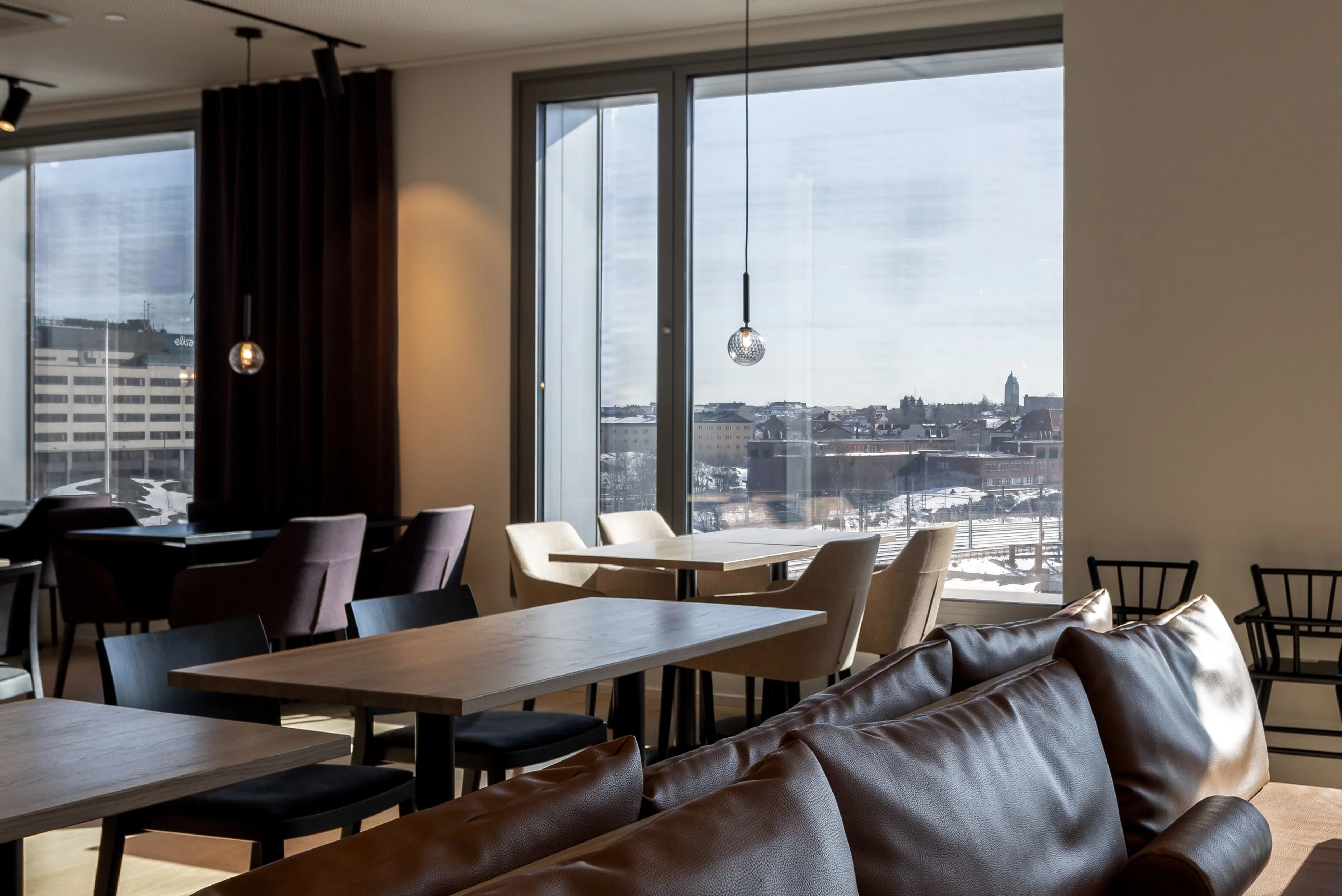 Moderni kahvila tai ravintola, jossa on pöytiä ja tuoleja, suuret ikkunat ja näkymä talviseen kaupunkimaisemaan. Tasainen lattia, hyvä valaistus.