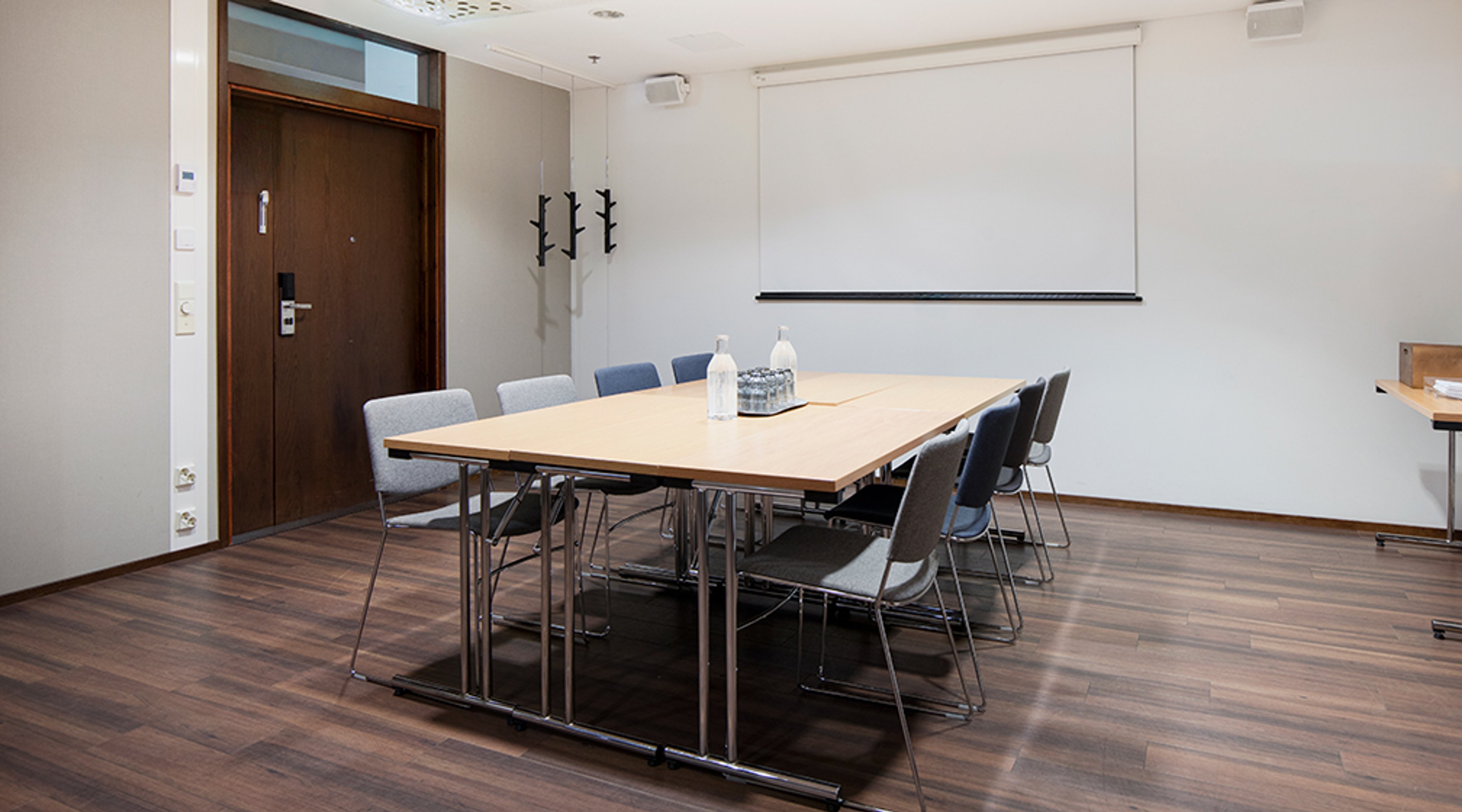 Neuvotteluhuone, jossa puinen pöytä kahdeksalla harmaaverhoilulla tuolilla, valkokangas seinällä ja tumma puulattia.