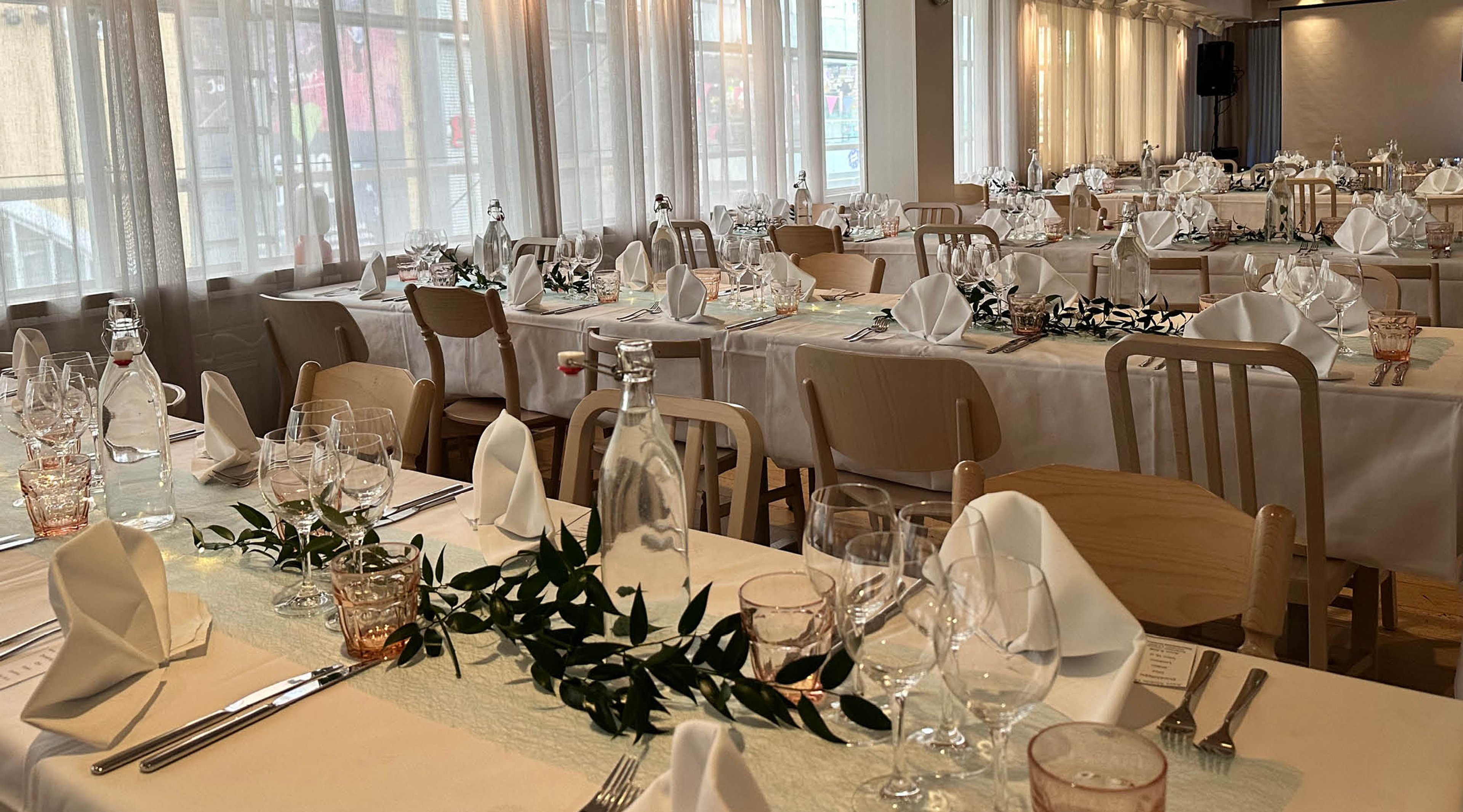 Juhlatila, jossa on useita pöytiä katettuna valkoisilla liinoilla. Puiset tuolit, viinilasit, servetit ja vihreät lehtikoristeet pöydillä. Isot ikkunat tuovat valoa tilaan.